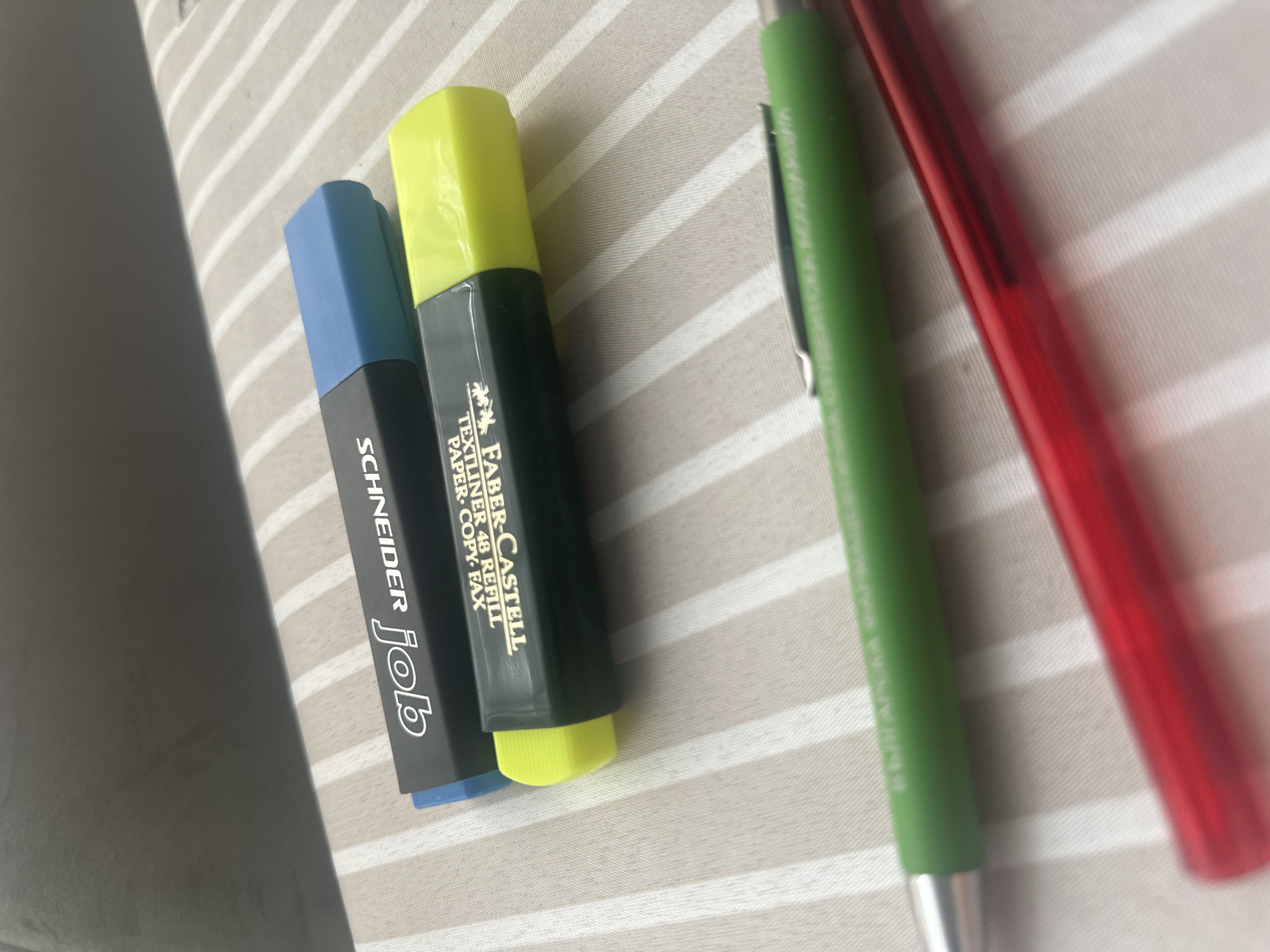 The image shows a close-up of three writing instruments lying on a surface with light grey and white stripes. From left to right, there is a blue-topped, black-bodied "Schneider Job" highlighter, a yellow-topped "Faber-Castell Textliner 48 Refill Paper Copy Fax" highlighter, and a green pen, partially obscured by a red pen. The location in the city of Sofia, Bulgaria cannot be determined from the image.