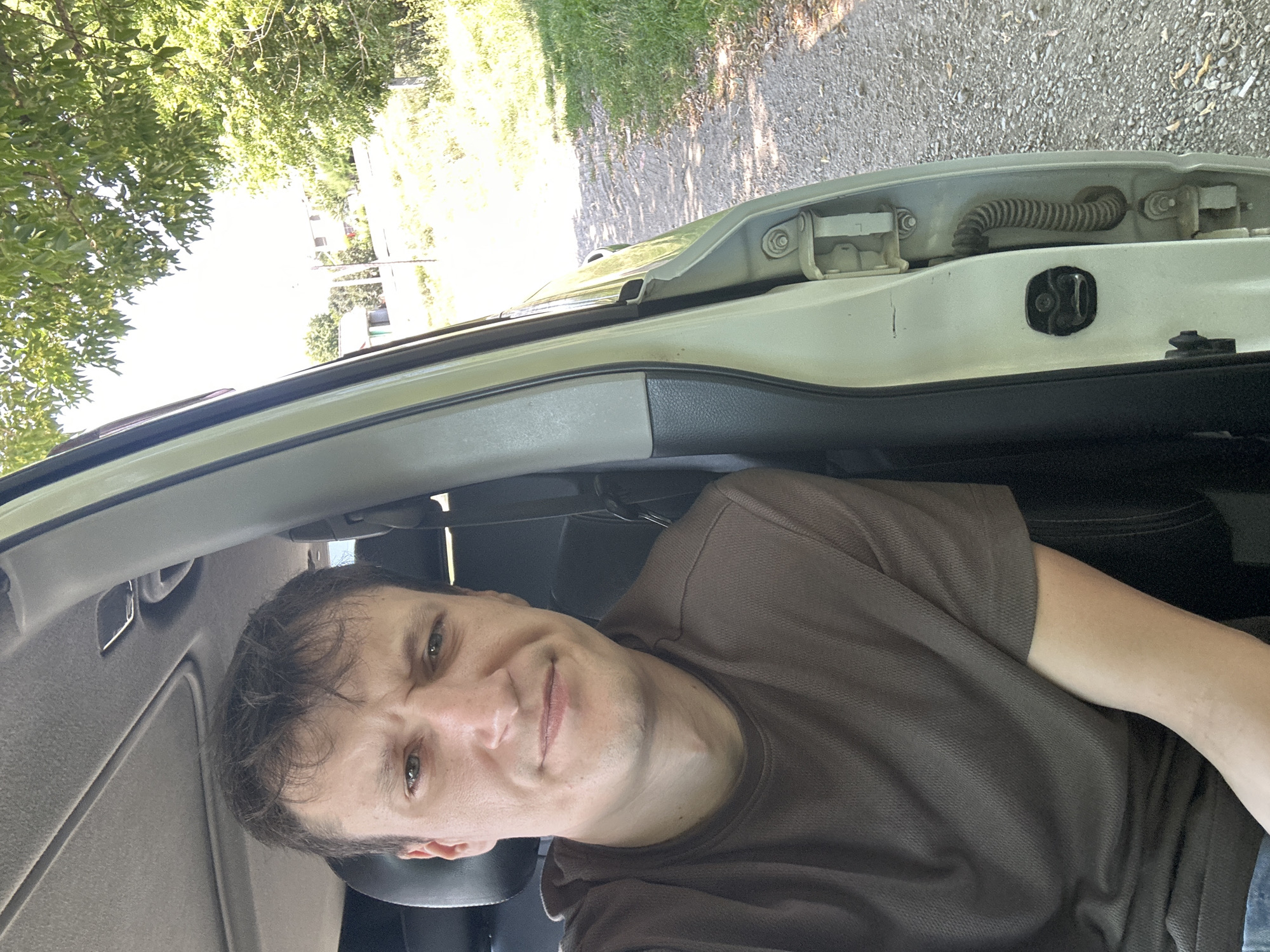 A man is sitting in the driver's seat of a vehicle, with the driver's side door open. He is looking at the camera with a slight grimace. The vehicle appears to be parked on a gravel road, surrounded by greenery and trees. In the background, there are houses and a clear sky, suggesting a rural or suburban setting outside the city of Makiv, Ukraine. The man is wearing a brown t-shirt. The vehicle's interior is visible, including the car door frame and a portion of the seat. The lighting suggests it is daytime.