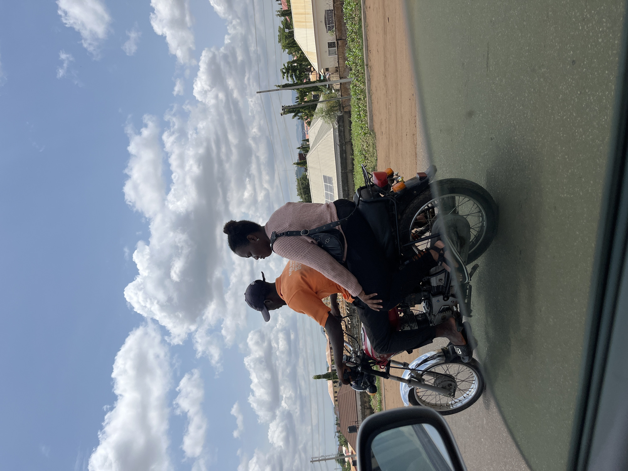 The image shows two people riding a motorcycle on a street in Abuja, Nigeria. The rider is a man wearing an orange t-shirt and a black cap. The passenger is a woman wearing a pink sweater and carrying a black bag. The motorcycle is on a paved road with a dirt shoulder. Buildings and vegetation are visible in the background. The sky is blue with scattered clouds. The image appears to have been taken from a car, as part of a car mirror is visible in the bottom left corner.
