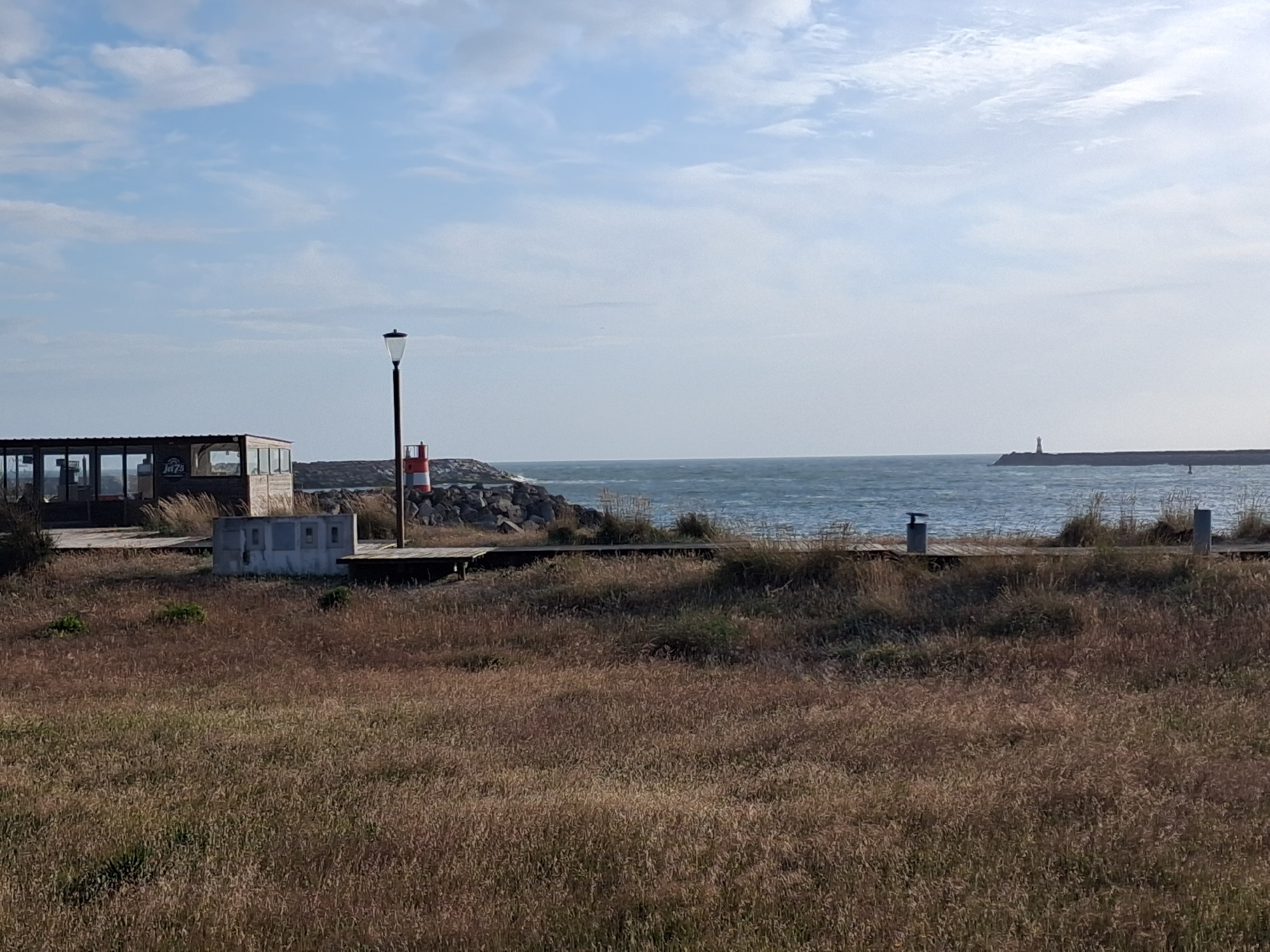 The image depicts a coastal scene in Figueira da Foz, Portugal. In the foreground is a field of dry, brown grass. Behind the grass, a wooden boardwalk runs along the coast. Several structures are visible along the boardwalk including a small, enclosed building with windows, a lamppost, and a concession stand. Beyond the boardwalk and structures, there is a rocky breakwater with a red and white marker. In the background, the ocean stretches to the horizon, with a distant structure visible in the distance. The sky is partially cloudy with blue patches.