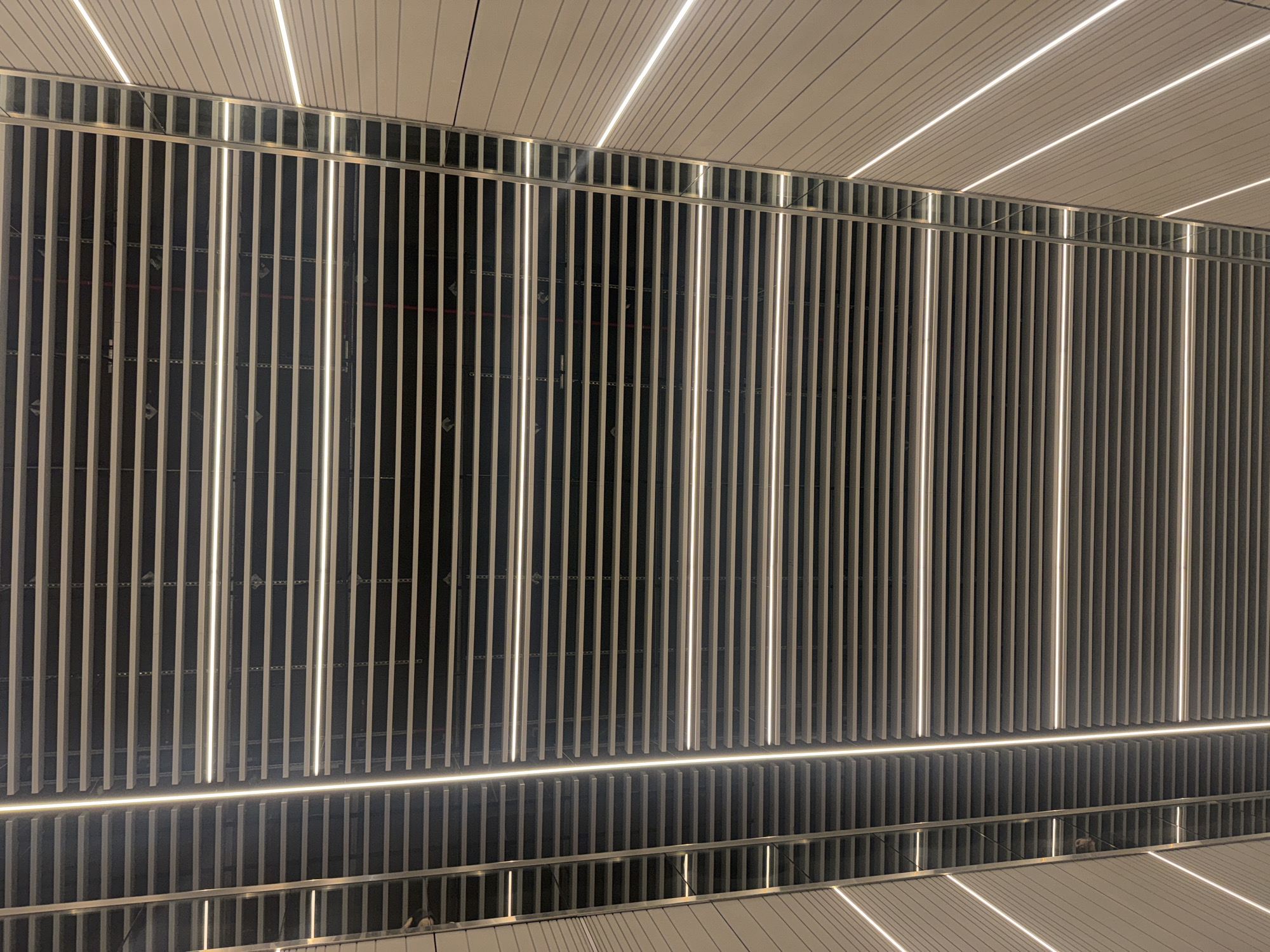 The image shows an architectural detail of a modern building, likely taken indoors. The primary elements are vertical slats or bars, probably part of a screen or wall. Interspersed among them are vertical light strips. Above the slatted structure is a ceiling composed of parallel lines, possibly panels or lights. A mirrored or reflective surface is at the bottom, creating a symmetrical, inverted reflection of the slatted structure and light strips. A small portion of a person can be seen as a reflection. This photo was taken in Ramat Gan, Israel.