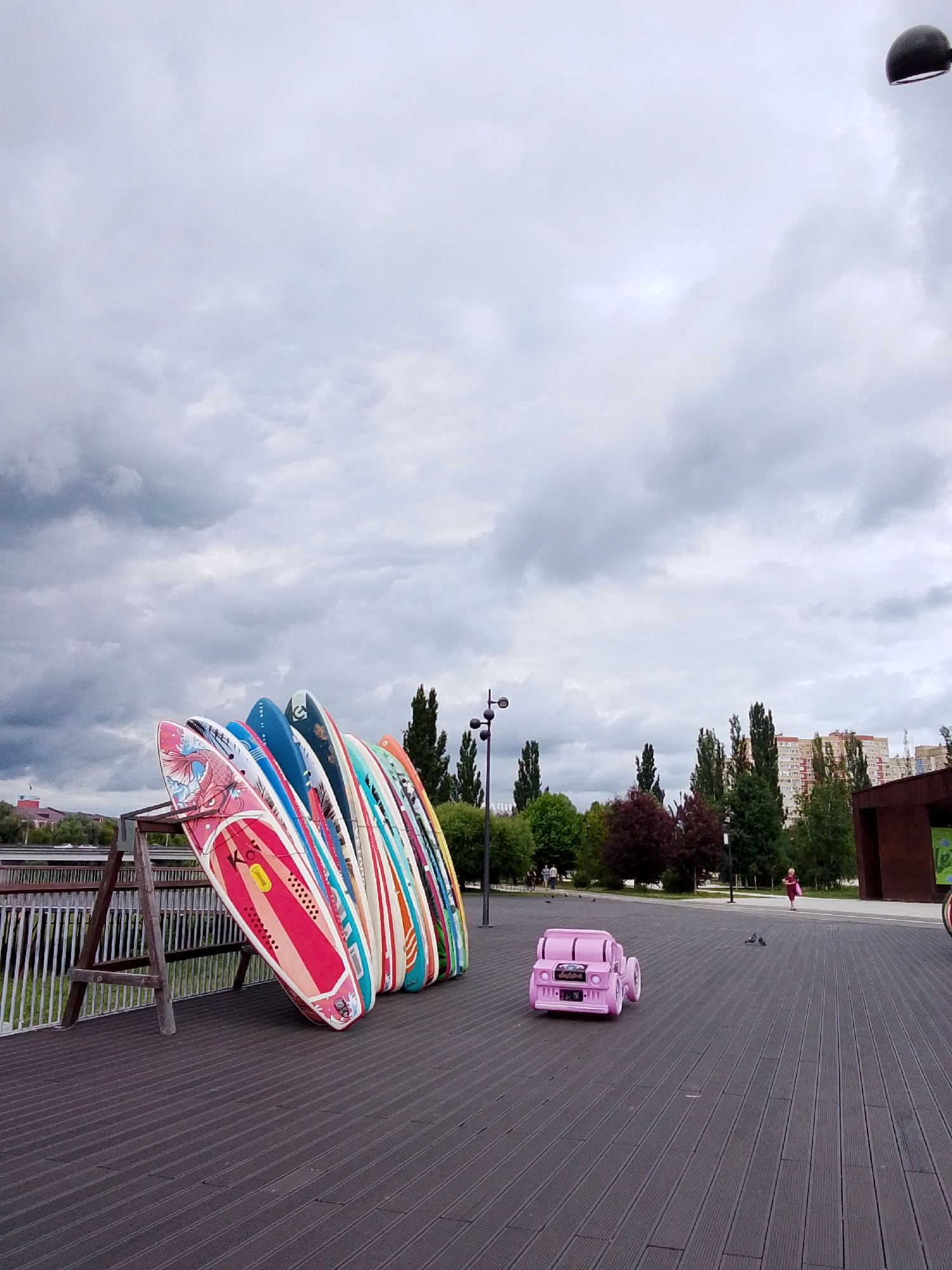 A group of colorful paddleboards are propped up on a wooden stand on a dark wooden boardwalk. In the distance, a small pink toy car sits on the boardwalk. A person in a pink dress walks away from the toy car towards a building in the background. The sky is overcast with gray clouds. Trees and buildings are visible in the distance. The scene appears to be a public park or waterfront area.