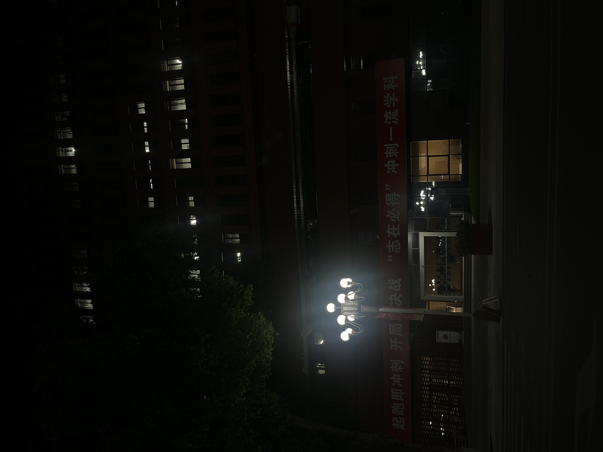 The image depicts a multi-story building at night in Yuzhong District, China, illuminated by internal and external artificial light sources. The primary structure is a dark-toned building, possibly brick or concrete, with numerous rectangular windows on its upper floors, several of which show interior lighting. The lowest floor features a prominent glass entrance with visible interior lighting and additional external lighting fixtures.
Across the lower facade of the building, a red banner with white Chinese characters is displayed. The characters translate to: "Hit the ground running, decisive battle, 'determined to win,' sprint towards a first-class discipline/subject."
In the mid-ground, to the left of the building's center, stands a street lamp with nine glowing spherical luminaires mounted on a decorative post. A vertical signpost is affixed to the street lamp's pole, with an A-frame sign positioned at its base on the ground. To the left of the street lamp, a large tree with dark, dense foliage partially obscures the upper-left section of the building.
The foreground consists of a dark asphalt street, showing faint horizontal markings, possibly indicating a pedestrian crossing. A paved area, likely a sidewalk, is visible directly in front of the building's entrance. No individuals are discernible in the scene. The overall lighting is low, with illumination concentrated around the building's windows, entrance, and the prominent street lamp.