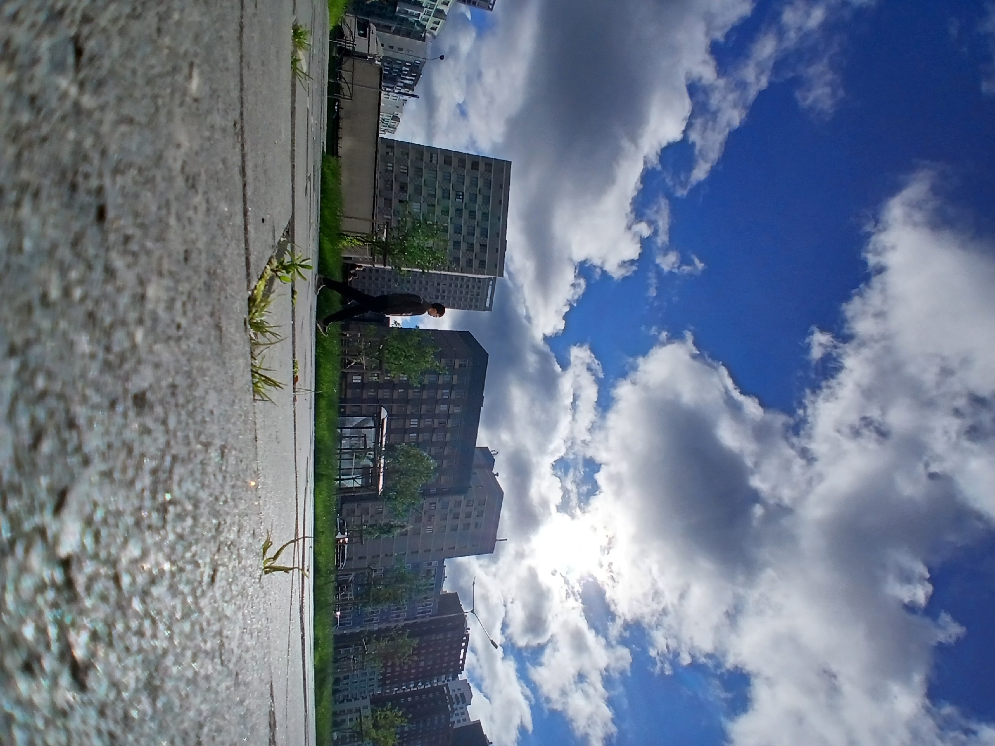 A man is walking on a paved path in an urban environment. The path is made of large grey pavers, with grass growing in the cracks. In the background, there are several multi-story buildings, some with balconies and many windows. Trees and a bus stop are visible between the path and the buildings. The sky is blue with scattered white and gray clouds. The sun is visible behind a cloud, creating a bright spot in the sky. The image appears to be taken from a low angle, emphasizing the foreground paving stones and the sky. Based on the architectural style of the buildings and the presence of a truck with Russian license plates visible on the left side of the frame, this scene is likely located in Yekaterinburg, Russia, possibly in a residential or commercial district.
