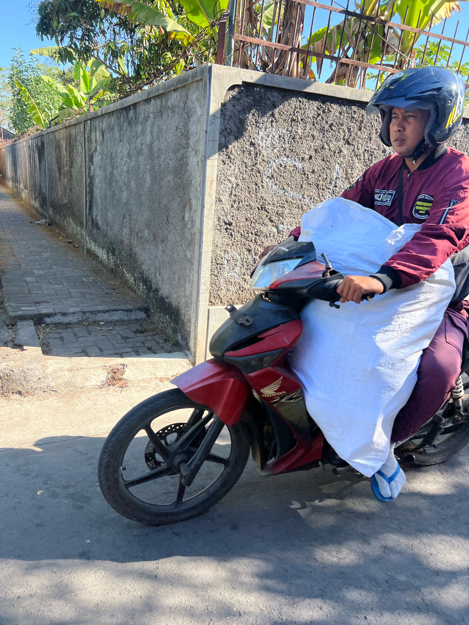 A man rides a red and black Honda motorcycle on a paved road, carrying a large white sack in front of him. He wears a maroon jacket with patches on the sleeve, a black helmet with yellow accents, and maroon pants. His right foot is clad in a blue flip-flop. The motorcycle has a visible "Honda" logo. The background features a textured gray concrete wall with dark staining, a brick pathway to the left, and lush green foliage and a metal fence at the top. The scene appears to be in a residential area in Payaman, Indonesia, with sunlight casting shadows on the road.