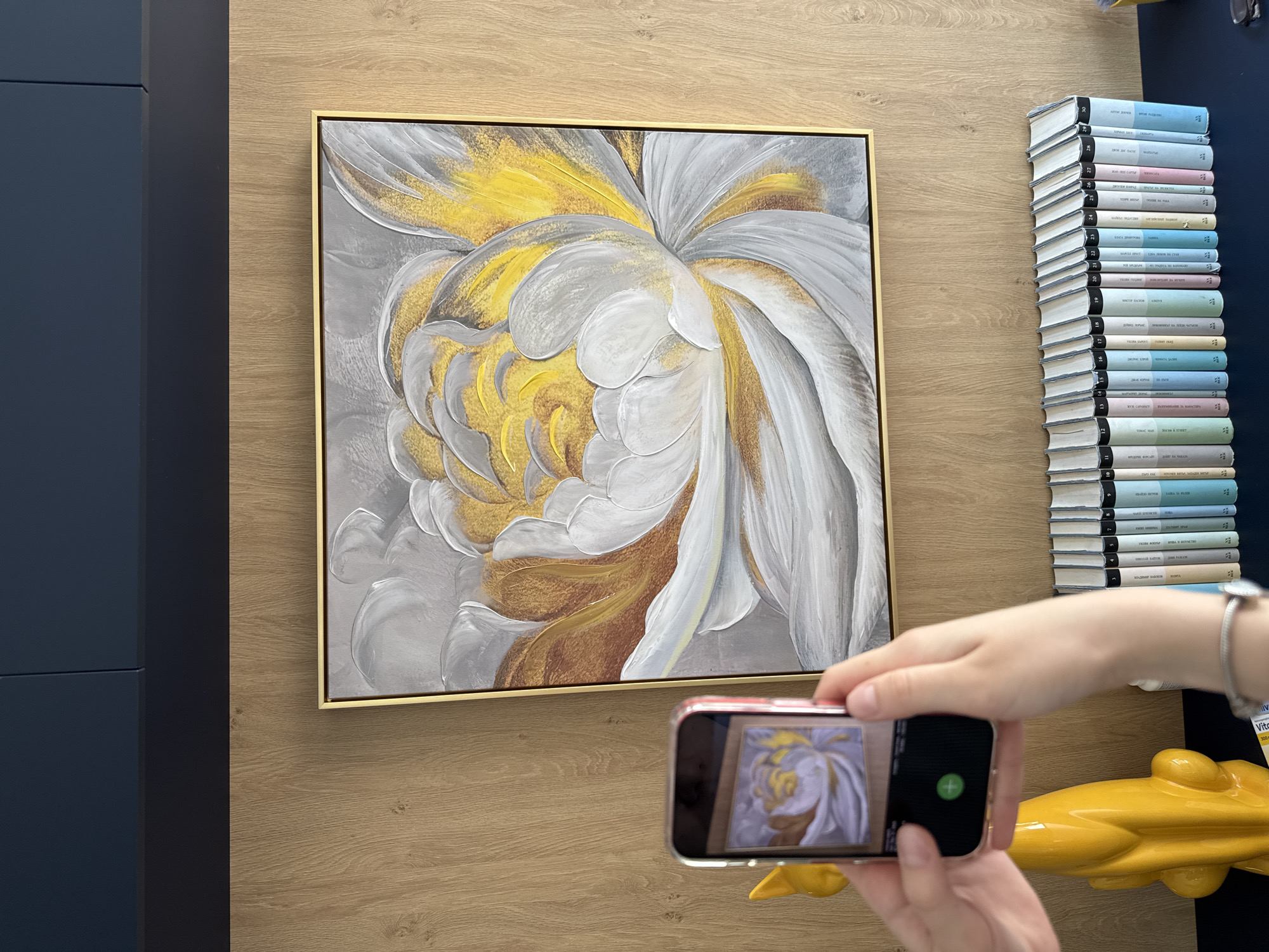 A person is taking a picture of a framed painting of a flower with a smartphone. The painting is hanging on a wall with a wood-grain pattern. To the left of the painting is a dark blue cabinet. To the right of the painting is a stack of books and a yellow object.
The location is likely in Sofia, Bulgaria, due to the Cyrillic lettering on the spines of the books.
