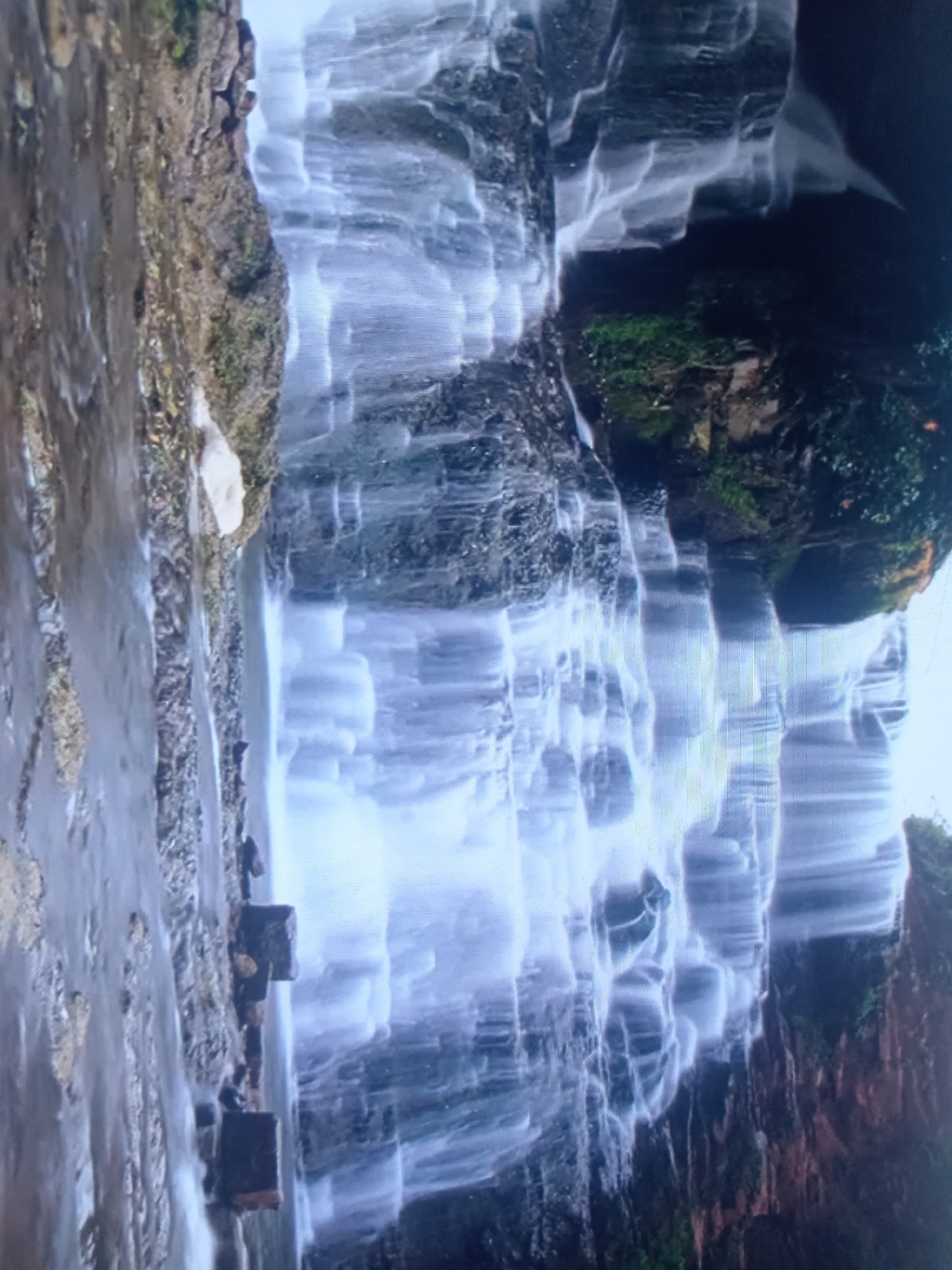 The image shows a multi-tiered waterfall cascading over moss-covered rocks. The water appears misty and diffused due to a long exposure, creating a soft, ethereal effect. The surrounding environment is lush and green, with vegetation clinging to the rocky cliffs. In the foreground, the water flows over a series of ledges, with some darker, rectangular objects visible on the left side, potentially man-made structures or natural rock formations. The overall impression is one of natural beauty and tranquility. The image is oriented vertically, suggesting a portrait perspective of the waterfall.