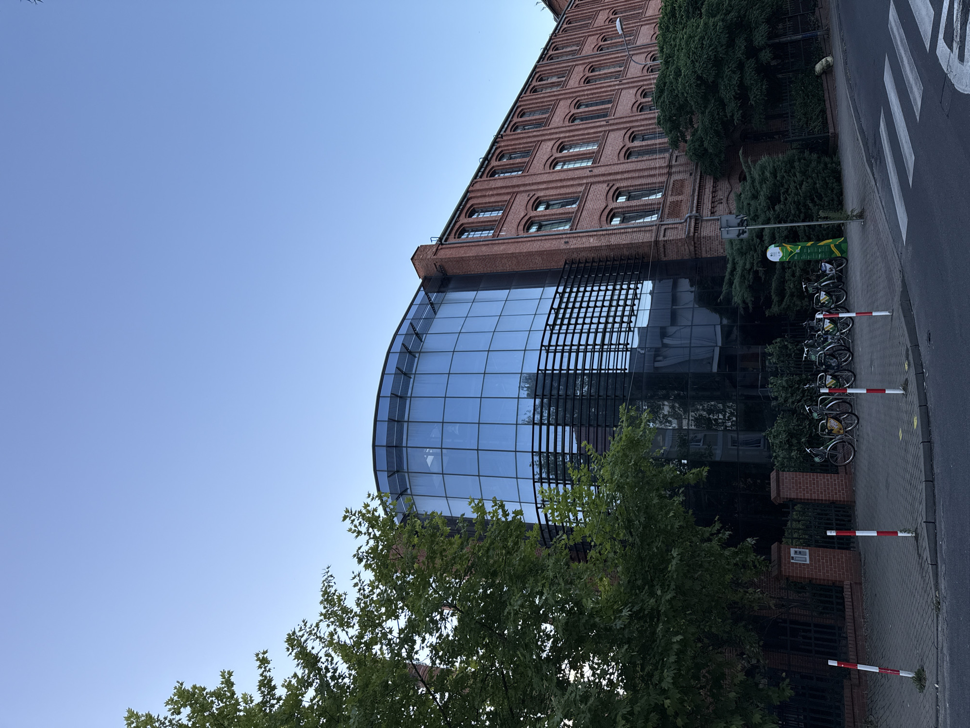 The image shows a multi-story building with a brick facade and a curved glass section. The building appears to be an office building or a repurposed industrial structure, possibly located in the city of Zielona Góra, Poland, given the architectural style which is reminiscent of former industrial areas in Polish cities. In front of the building, there is a sidewalk with several bicycles parked. A crosswalk is visible on the street. Trees and green bushes surround the building and the sidewalk. The sky is clear and blue. The overall scene suggests an urban environment, likely during daytime.