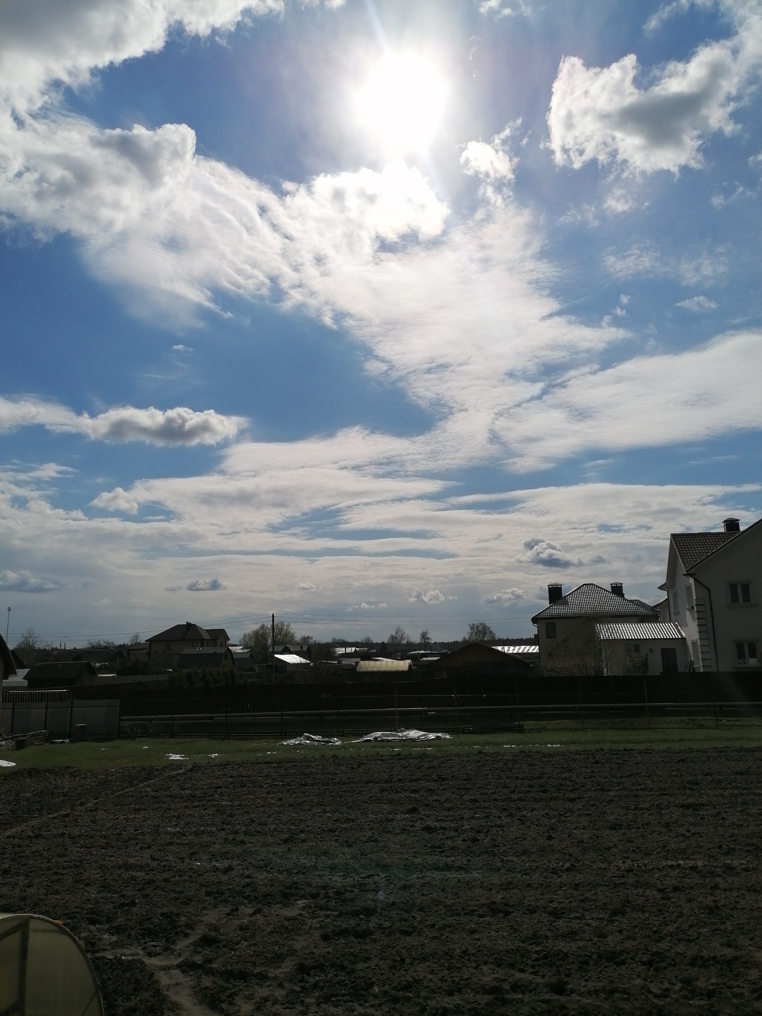 The image presents a bright outdoor scene, likely a rural or suburban landscape during late winter or early spring. The upper portion of the frame is dominated by a vivid blue sky, filled with an array of clouds varying from delicate, wispy cirrus streaks to more substantial, fluffy cumulus formations. The sun shines intensely from the upper right, casting a brilliant light and creating a noticeable lens flare, indicating a clear and sunny day.
In the middle ground, a line of detached houses with pitched, dark-tiled roofs stretches across the horizon. Some of these residential buildings feature light-colored facades, while others appear darker. Interspersed among the houses are trees, mostly bare or just beginning to show signs of early foliage. Various fences and boundaries are visible, separating the properties.
The foreground is composed primarily of a large, dark expanse of disturbed earth, possibly a tilled field or muddy ground, suggesting recent agricultural activity or preparation for planting. Beyond this dark field, a strip of green grass is visible, containing small, scattered patches of white that resemble remaining snow or ice, further reinforcing the seasonal context. In the very bottom-left corner, a small, translucent, arch-shaped structure, possibly part of a greenhouse, is partially visible. There are no people or explicit activities depicted, and no visible text. The overall atmosphere is one of natural light and transition between seasons.