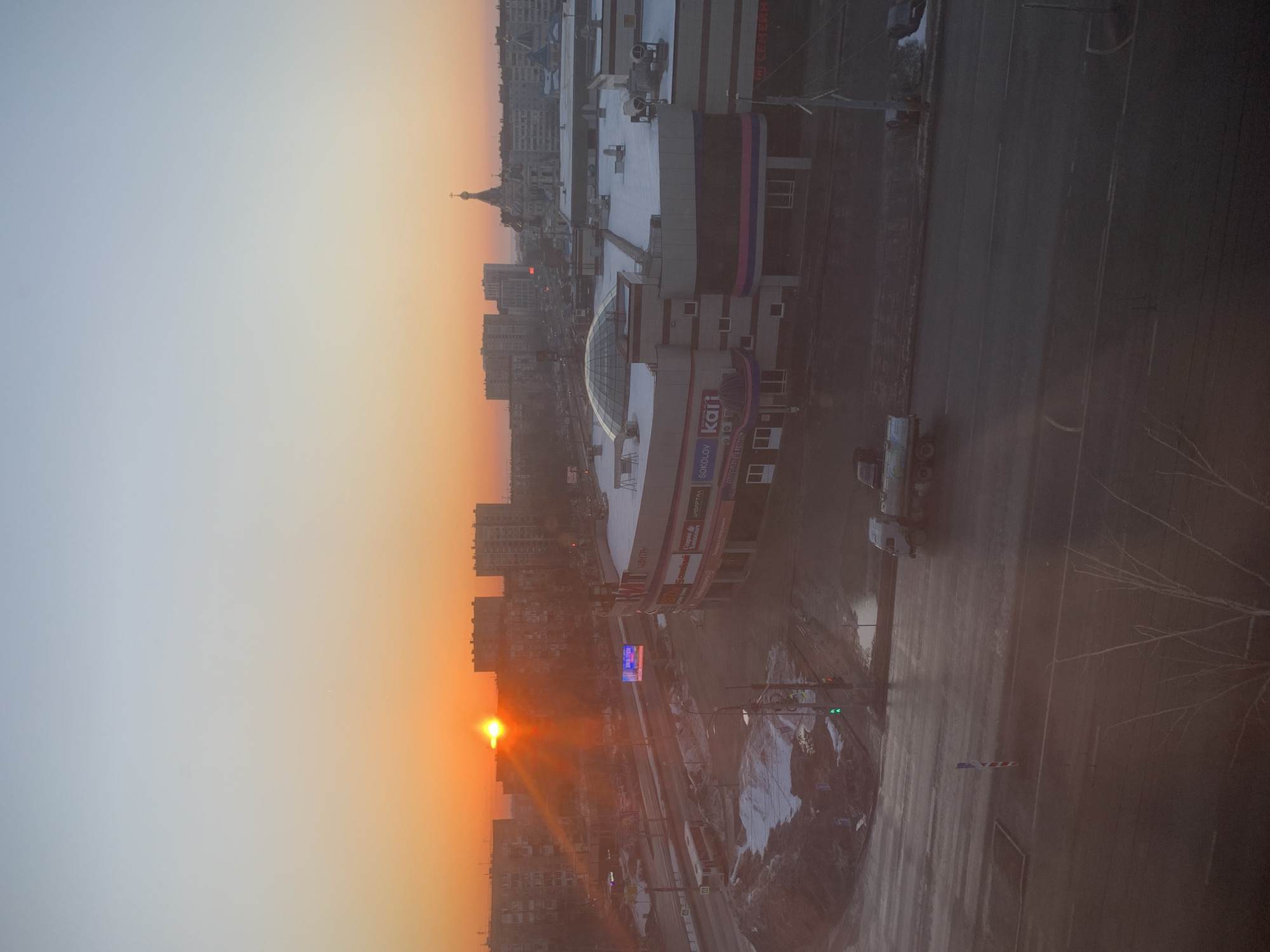 An elevated, wide-angle view captured during sunrise depicts a city scene in Izhevsk, Russia, specifically showing the area surrounding the "ТРЦ Столица" (TRC Stolitsa) shopping mall and St. Michael's Cathedral. The sun, a bright orange disc, is low on the horizon on the far left, casting strong orange light and lens flare, illuminating the lower sky in hues of orange and fading to light blue overhead.
In the mid-ground, the multi-story "ТРЦ Столица" shopping mall is prominent on the right. Its roof is partially covered in snow, featuring a large glass dome structure. Visible storefront signs on the mall's facade include "SOKOLOV" and "kari", along with Cyrillic text such as "Пятерочка" (Pyaterochka) and "Центр". The building exhibits a modern design with horizontal pink, purple, and blue striped accents.
To the right and slightly behind the mall, the golden domes and spire of St. Michael's Cathedral are visible, rising above the surrounding cityscape. In the background, numerous multi-story residential buildings, consistent with Soviet-era architecture, stretch across the horizon.
A multi-lane asphalt road runs horizontally across the lower-mid section of the image, appearing wet, with puddles reflecting the dim light. Snow piles are present along the road's edges and in undeveloped ground areas. A white tanker truck is visible on the road in the lower-middle, facing left. Further left, a white bus or trolleybus is present on the road. A green traffic light is visible near the bottom left corner. An illuminated advertising screen, displaying blue and white content, is positioned across the street from the shopping mall. Bare tree branches are visible in the immediate foreground on the lower left.