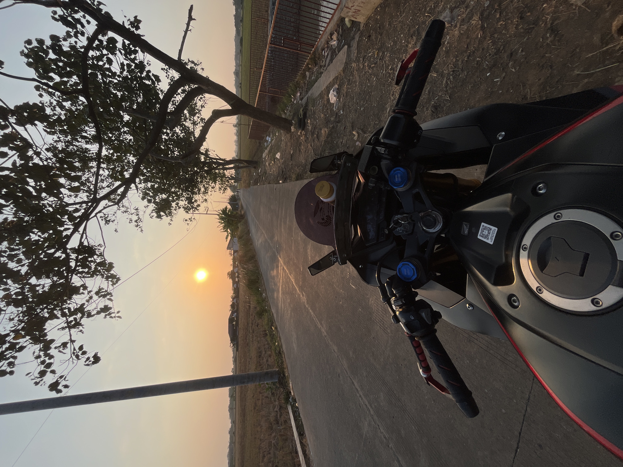 The image captures a first-person perspective of a motorcycle parked on the side of a road, likely in the city of Pecangaan, Indonesia, during sunset. The road is paved, with dried rice fields on the left and a canal with a fence on the right. A tall, slender utility pole stands to the left of the road, and trees with sparse foliage line the right side. The sun is low on the horizon, casting a warm, golden glow over the scene. The motorcycle's handlebars, front brake, throttle, and part of the fuel tank are visible in the foreground. A small brown bottle with a yellow cap is perched on the motorcycle's dashboard. The sky is clear with a gradient of orange and blue. The overall atmosphere is serene and rural.