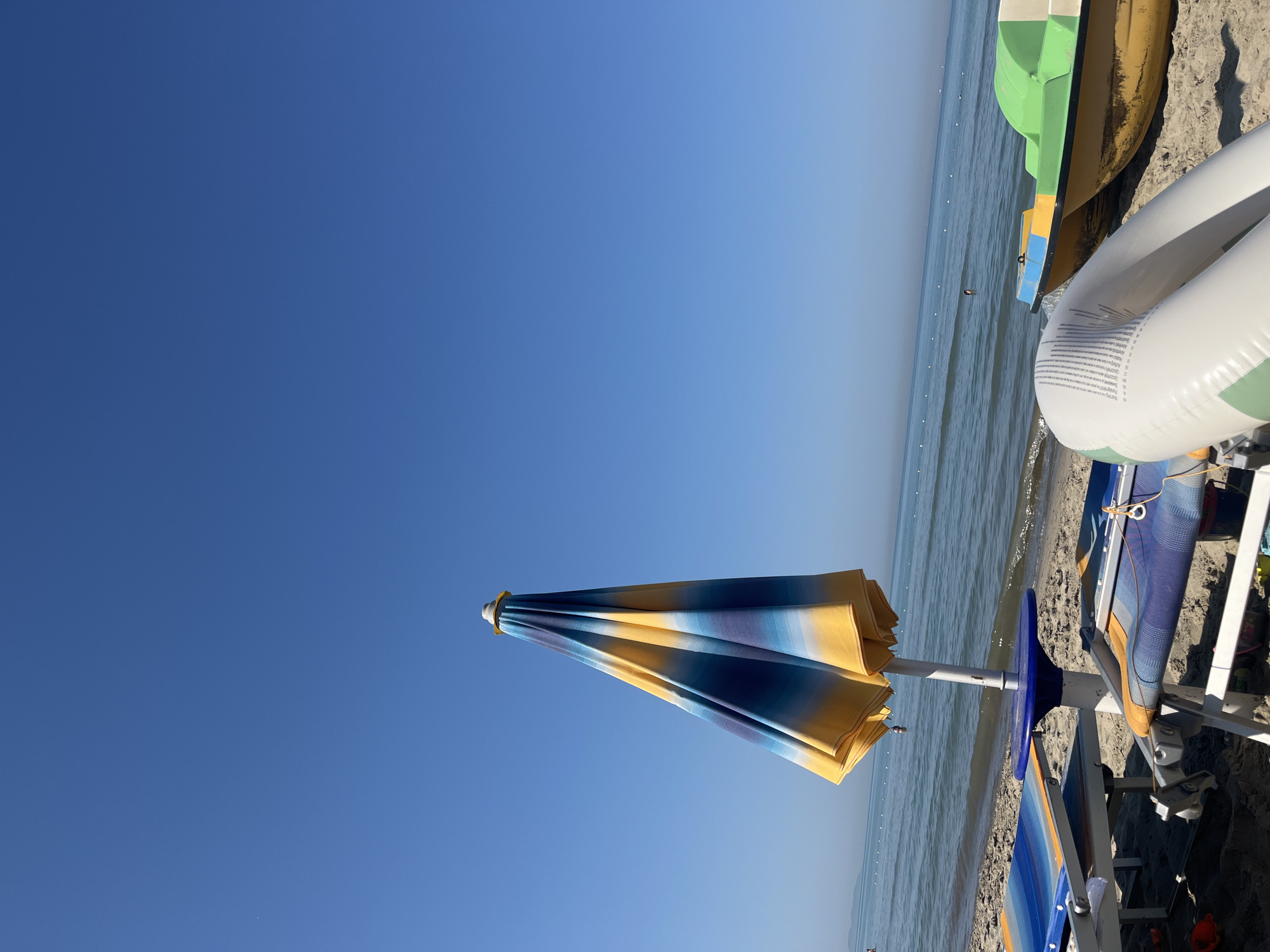 The image depicts a beach scene in Bashkia Durrës, Albania, under a clear blue sky. In the foreground, two beach chairs with blue and orange striped upholstery are set up on the sand. A closed, colorful beach umbrella with yellow, blue, and white stripes stands beside them. To the right, a yellow and green paddleboat is partially visible on the sand, with a white inflatable tube in front of it. The calm sea stretches towards the horizon, dotted with a few distant buoys and a solitary swimmer. The sandy beach slopes gently towards the water.