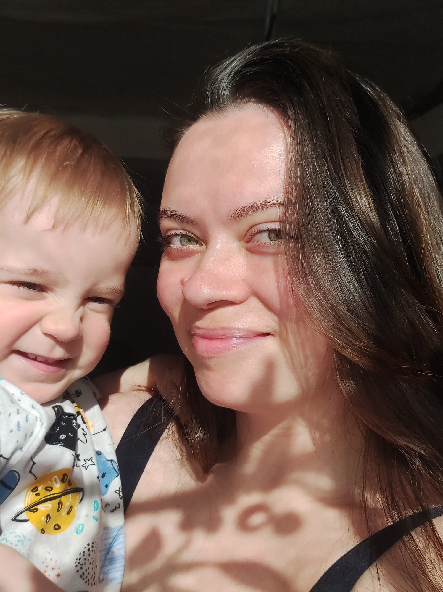 A selfie-style photograph depicts a fair-skinned woman and a young child in an indoor setting, with light casting shadows across their faces and bodies. The woman, with dark hair and green eyes, smiles at the camera. She has a mole on her right cheek. The child, with blond hair, is held close to the woman, his face exhibiting a gleeful expression. The child wears a white top printed with planets and cosmic objects. The woman is wearing a black tank top. The background is largely dark, suggesting a room or space away from direct light. The location is Unknown, Unknown.