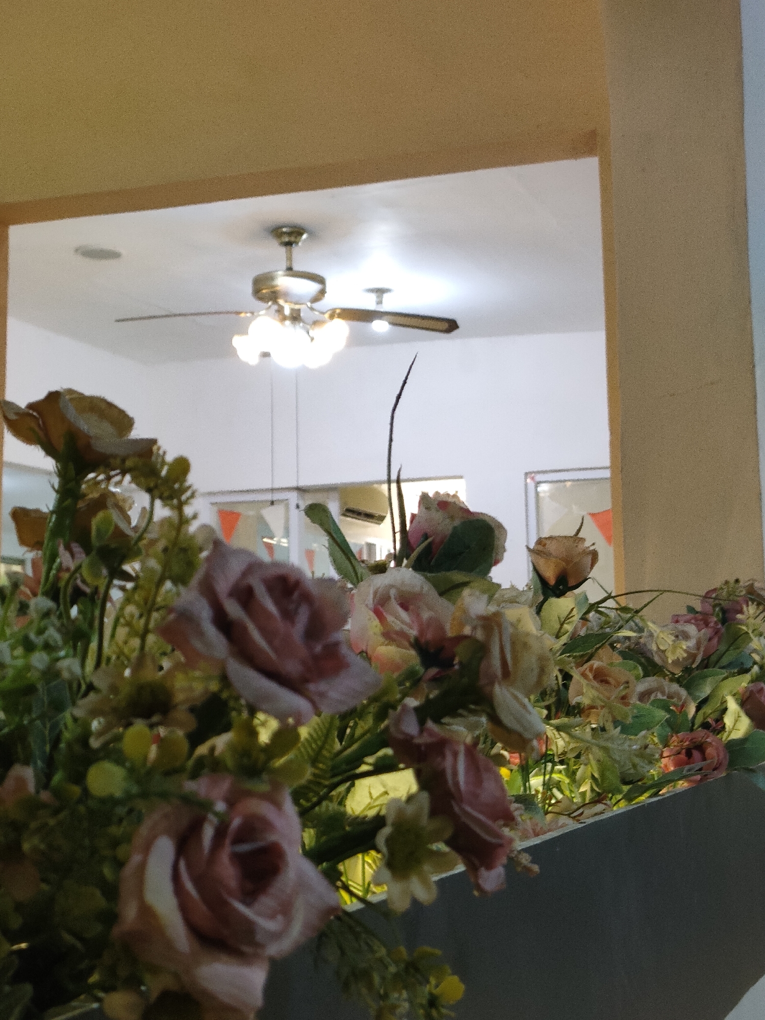 A close-up, low-angle shot captures a floral arrangement in the foreground, with a bright, modern interior visible in the background. The arrangement features several muted-pink and beige roses with green foliage. The background shows a white ceiling with a brushed nickel ceiling fan with multiple lights, and an air conditioning unit mounted on the wall. The overall scene suggests an indoor public space, possibly a restaurant or hotel lobby in Semarang, Indonesia. The lighting is warm, with artificial lights from the ceiling fan illuminating the area. The composition emphasizes the floral display, with the background slightly out of focus.