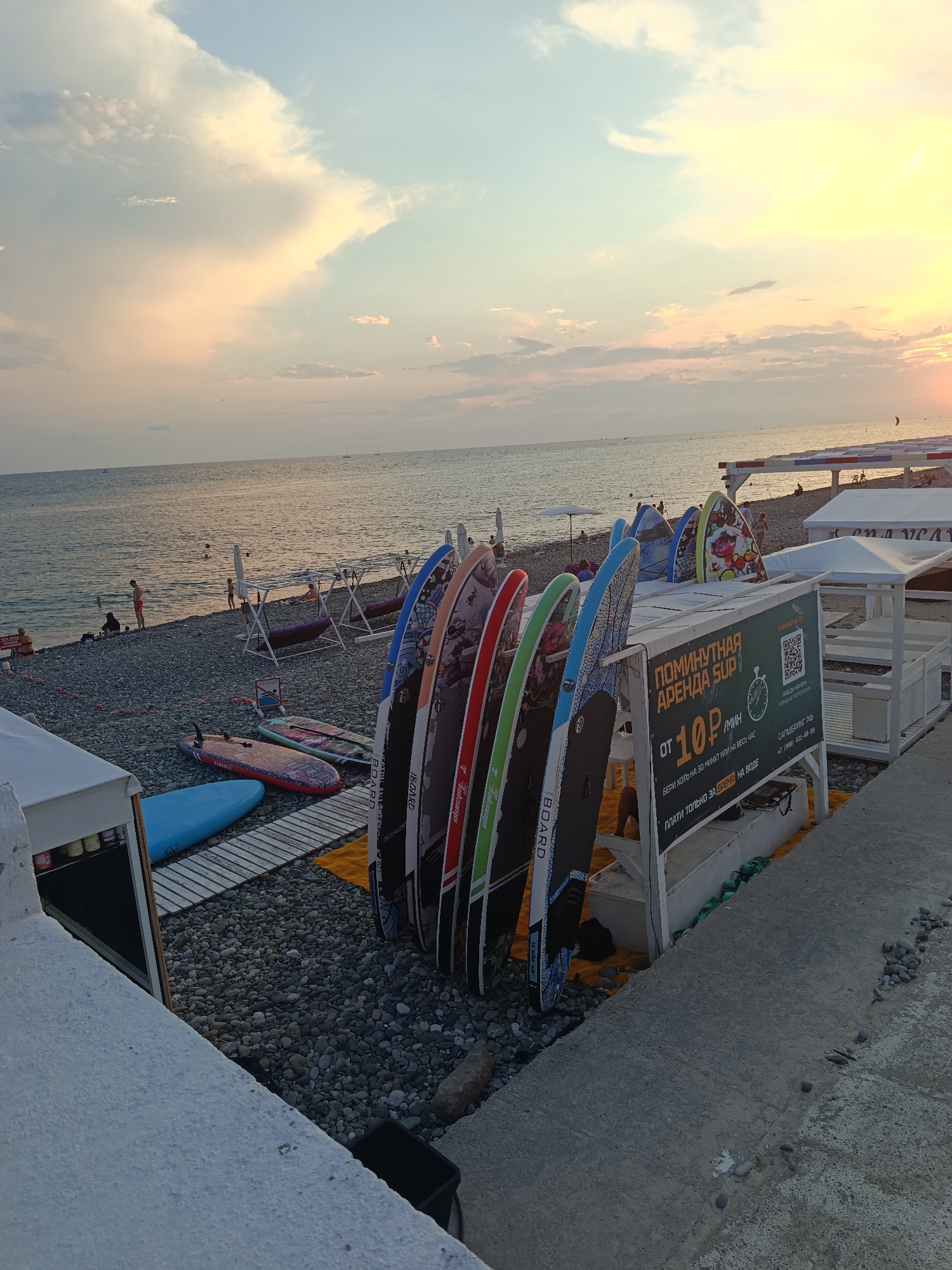 A wide shot captures a pebble beach in Sirius, Russia, at sunset. The sky is a gradient of soft blues, oranges, and yellows, with wispy clouds scattered across the horizon. The calm sea stretches out, reflecting the warm glow of the setting sun.
In the foreground, several stand-up paddleboards are neatly arranged vertically, leaning against a white structure that appears to be a rental stand. The boards are colorful and decorated with various patterns. A sign on the structure advertises "ПОМИНУТНАЯ АРЕНДА SUP" (Minute rental SUP) with a price of "10 ₽/мин" (10 rubles per minute), indicating a rental service for paddleboards. A QR code is also visible on the sign, likely for payment or information.
To the left of the paddleboards, other paddleboards are laid horizontally on wooden planks. A bright blue paddleboard is prominent. Further back, on the pebble beach, several people are visible. Some are wading in the shallow water, while others are standing or sitting on the beach. There are also some white beach umbrellas and lounge chairs, suggesting a recreational area. The overall scene conveys a peaceful and recreational atmosphere at the end of the day.