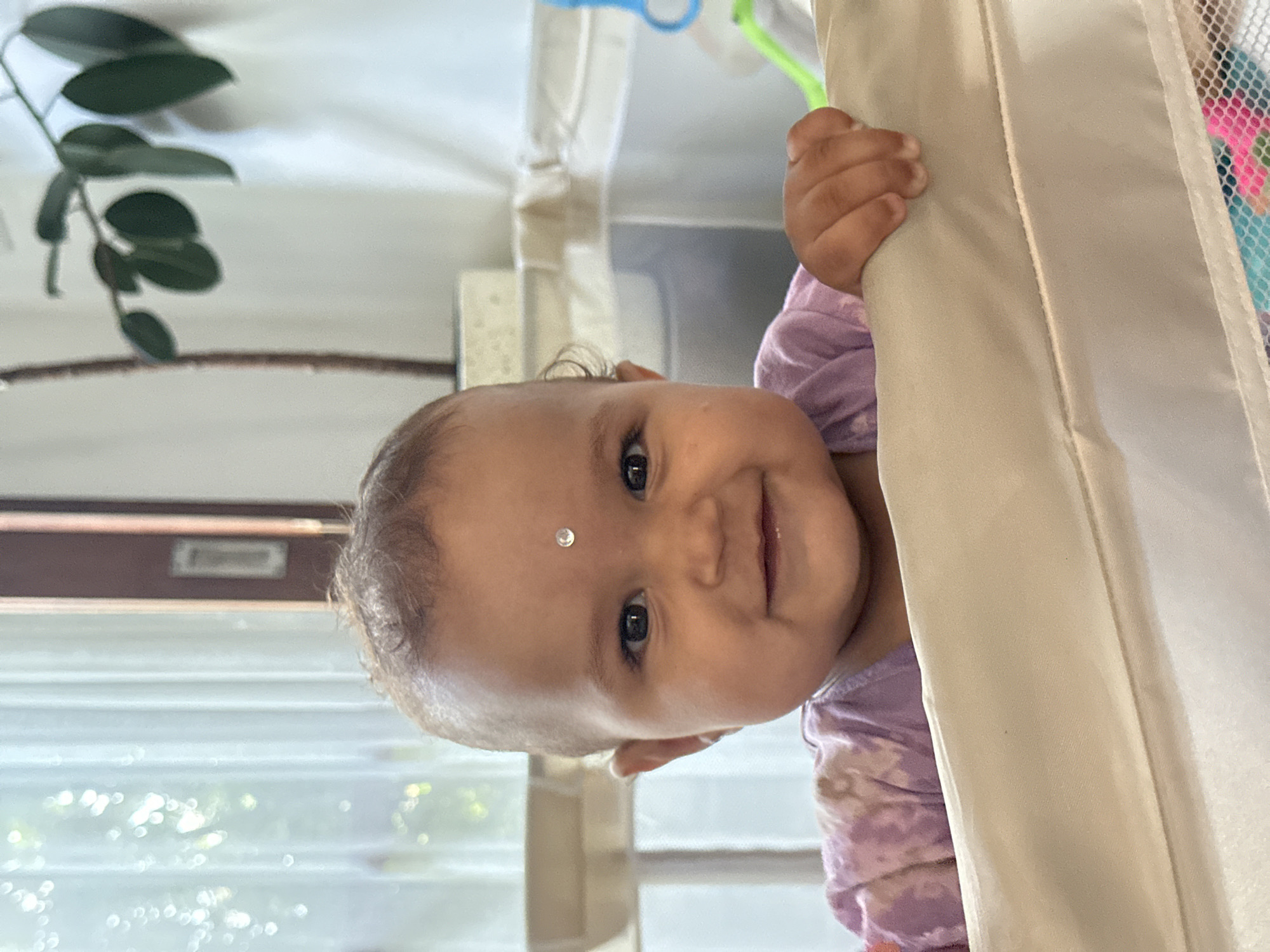 A baby with a circular silver sticker on its forehead is grasping a white fabric barrier and looking at the camera. The baby is wearing a lavender top. A window and green plant are visible in the background. The photo was taken indoors. The location is identified as North Kuta, Indonesia.