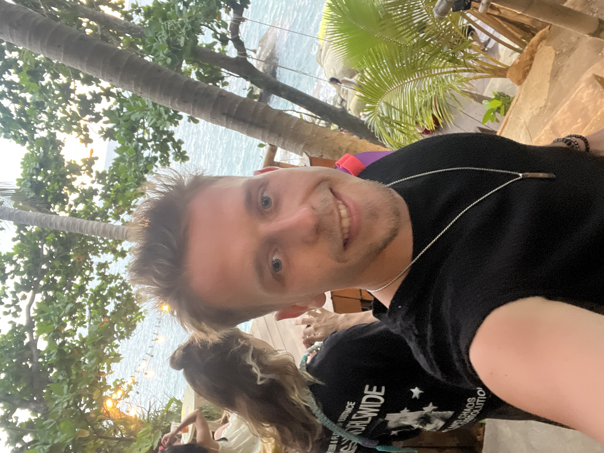 A man is taking a selfie in an outdoor setting, with a beach and ocean visible in the background. He is wearing a black t-shirt and a silver necklace. To his left, a woman with long brown hair is visible, wearing a black t-shirt with white text. She appears to be facing away from the camera and looking at something on her left. The environment suggests a tropical location, with palm trees and lush greenery. In the background, the ocean has gentle waves, and some rocks are visible near the shore. There are also string lights hanging between trees, suggesting a festive atmosphere or evening setting. The man's expression is a slight smile. The overall scene appears to be a casual gathering at a beachfront location. The clothing and vegetation indicate a warm climate, consistent with Karon, Thailand.