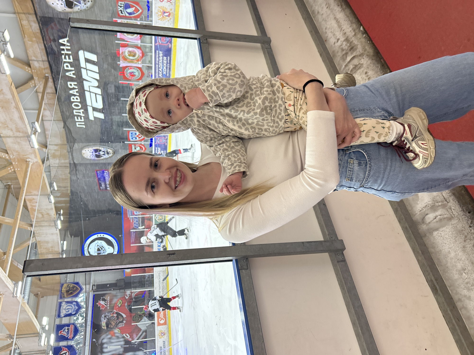 A woman holds a baby in front of a hockey rink at "Ledovaya Arena Temp" in Chaikovsky, Russia. The woman wears jeans and a white long-sleeved shirt. The baby wears a hooded jacket, pants, and shoes. The rink is visible through glass barriers, showing hockey players on the ice. Logos of various hockey organizations and teams are displayed in the background.