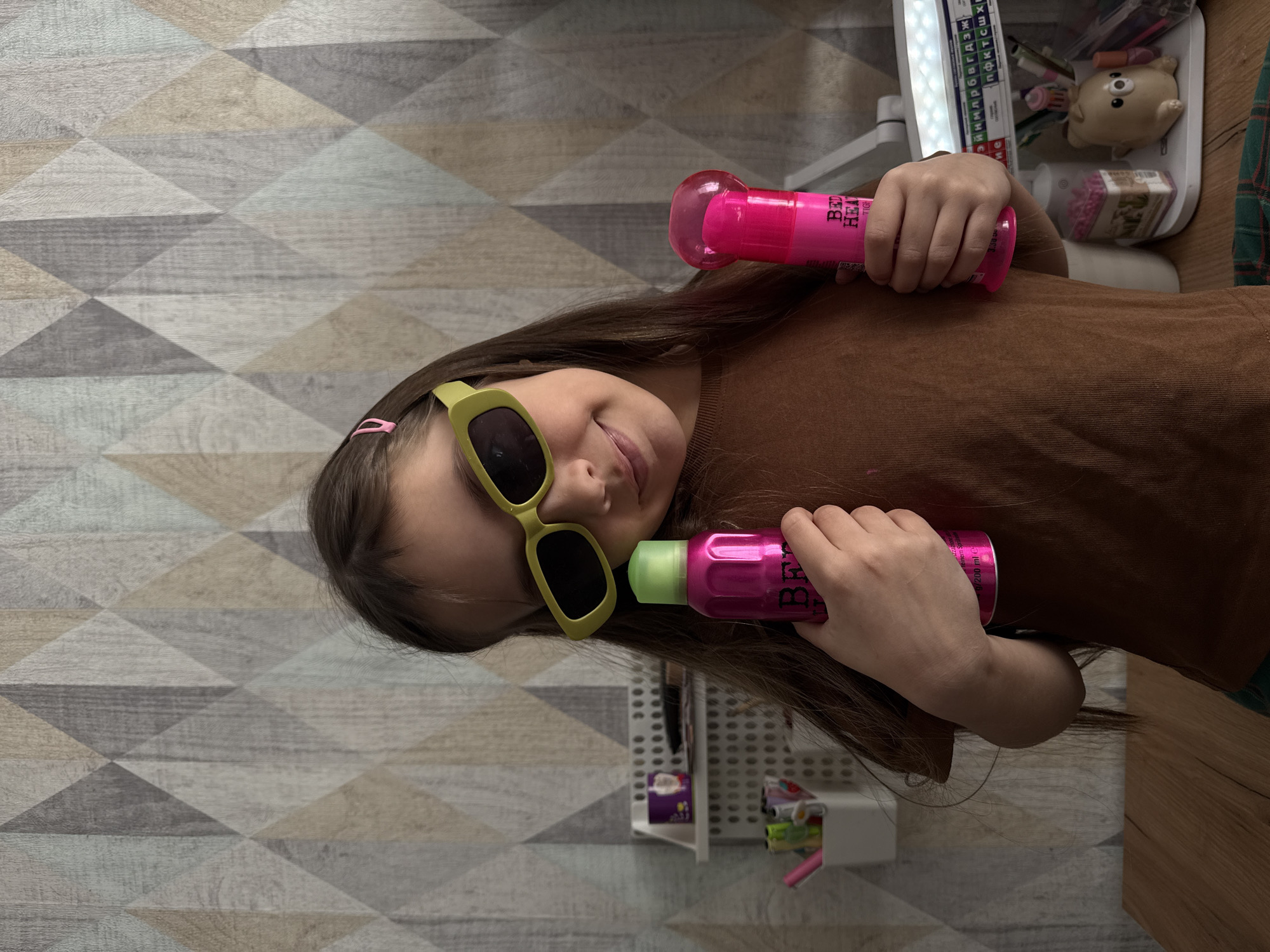 A young girl wearing yellow sunglasses and a brown shirt holds two pink spray cans. The background includes a geometric patterned floor and a shelf with various objects. The location is Votkinsk, Russia.