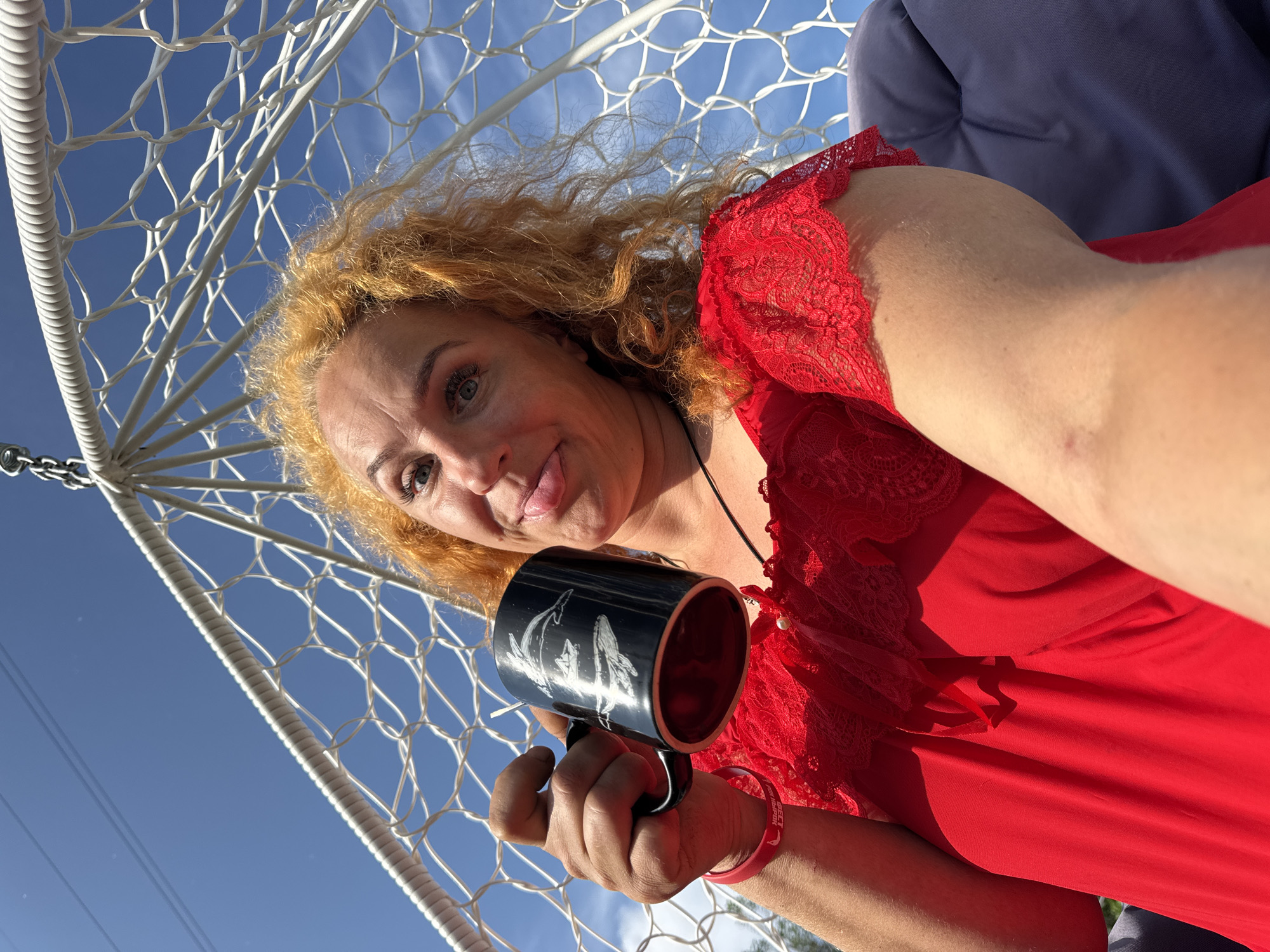 A woman with bright orange curly hair and blue eyes is sitting in a white hanging chair, holding a black mug with drawings of dolphins and whales. She is wearing a red dress with lace accents. The mug is decorated with white illustrations of marine life. The background is a clear blue sky with a few wispy clouds. The scene appears to be outdoors, possibly in a backyard or park setting, under bright sunlight. The woman is sticking her tongue out playfully at the camera. The image does not provide any specific information about the location in Chaikovsky, Russia, beyond the general outdoor setting.