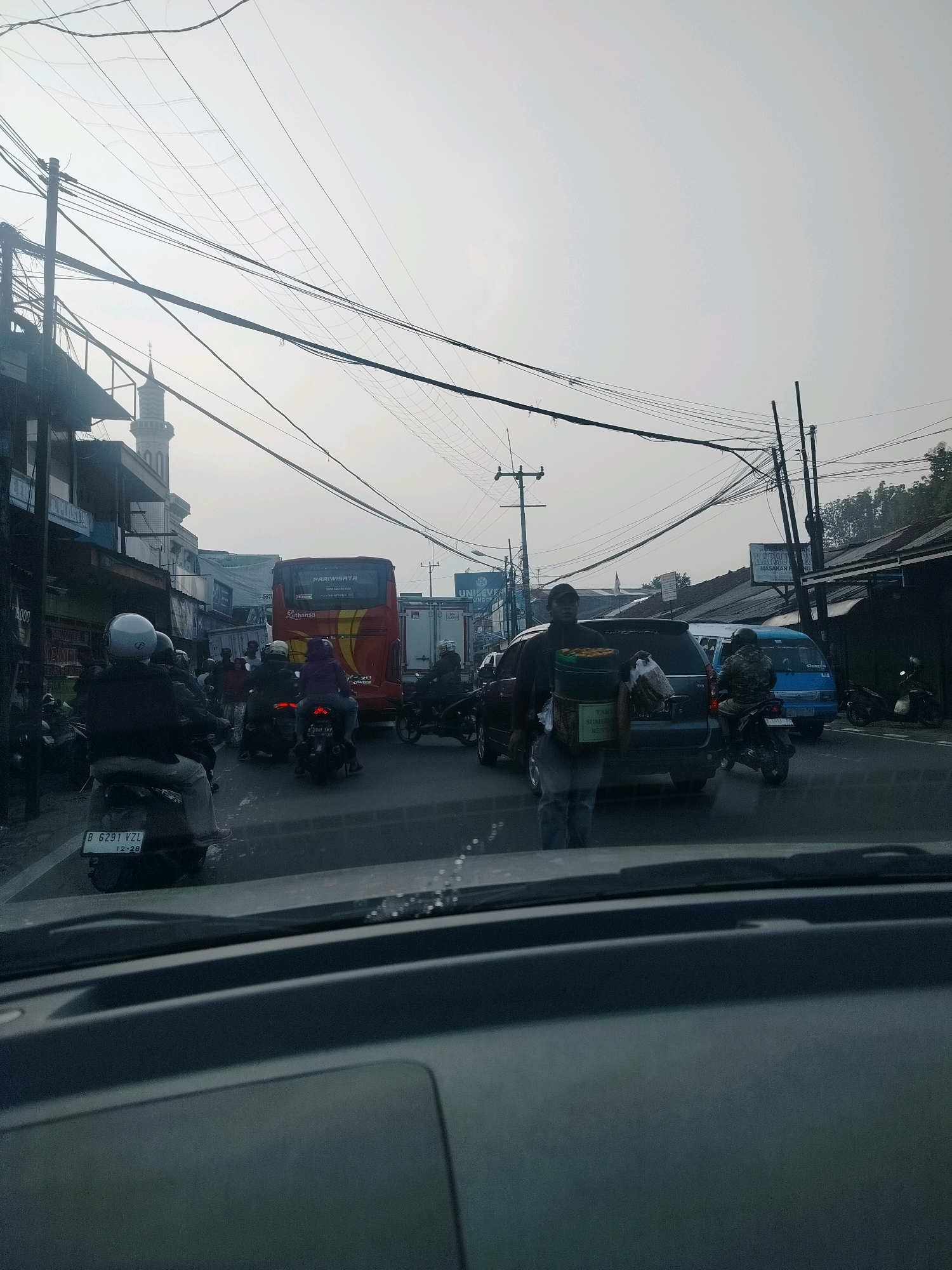 This image depicts a busy street scene in Megamendung, Indonesia, characterized by heavy traffic, overhead electrical wires, and buildings lining the road. A red tour bus, with "PARIWISATA" written on its side, is a prominent feature, stopped in traffic. Several motorcycles are also visible, with riders wearing helmets. A man is seen walking on the side of the road carrying a large container and a bag. The sky is overcast, suggesting a hazy or potentially polluted atmosphere common in urban settings. The foreground shows the interior of a vehicle, indicating the photo was taken from a car. The presence of a mosque minaret in the background further anchors the location within Indonesia.