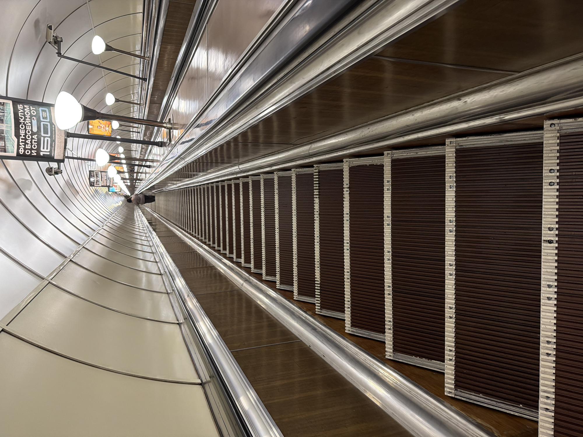 The image captures the escalator tunnel at the Admiralteyskaya metro station in Saint Petersburg, Russia. The tunnel is characterized by curved walls, lined with a series of evenly spaced globe-shaped lights. Advertisements are placed along the tunnel wall. A long escalator spans into the distance, its steps appearing progressively smaller due to perspective. The sides of the escalator showcase parallel handrails and decorative panels, adding to the tunnel's architectural depth.