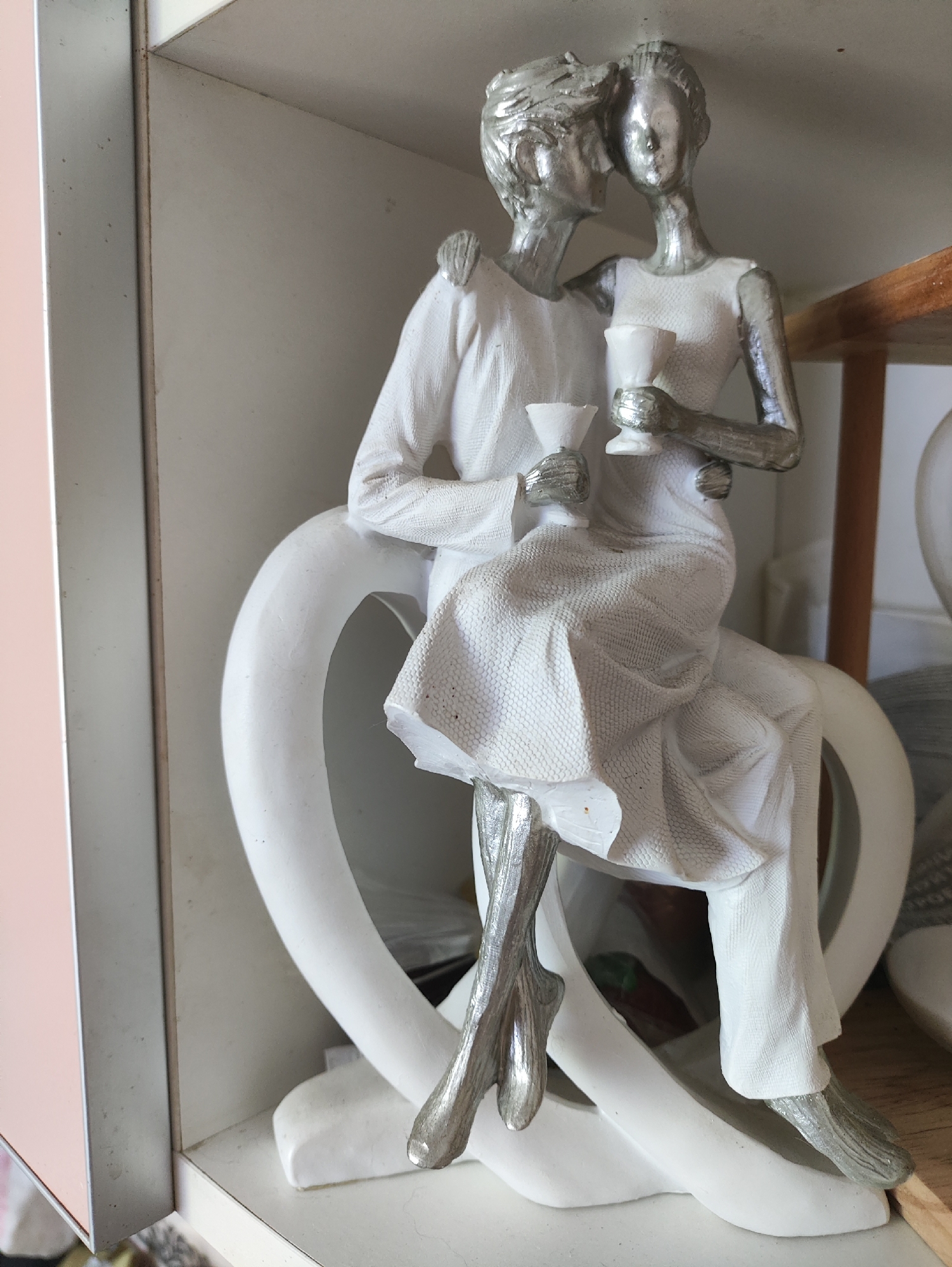 This is a close-up, eye-level shot of a decorative figurine of two individuals, likely a couple, seated and holding chalices. The figurine has a silver finish on the figures' bodies and clothing is rendered in white with textured fabric detail. They are positioned on a white, heart-shaped base. The figurines are depicted with their bodies close, one leaning in as if to kiss the other. The background consists of white shelving, with hints of other objects and a pale pink vertical panel on the left edge of the frame. The scene is indoors, likely in a domestic setting. There is no information available to determine the location in the city of Kharkiv, Ukraine.