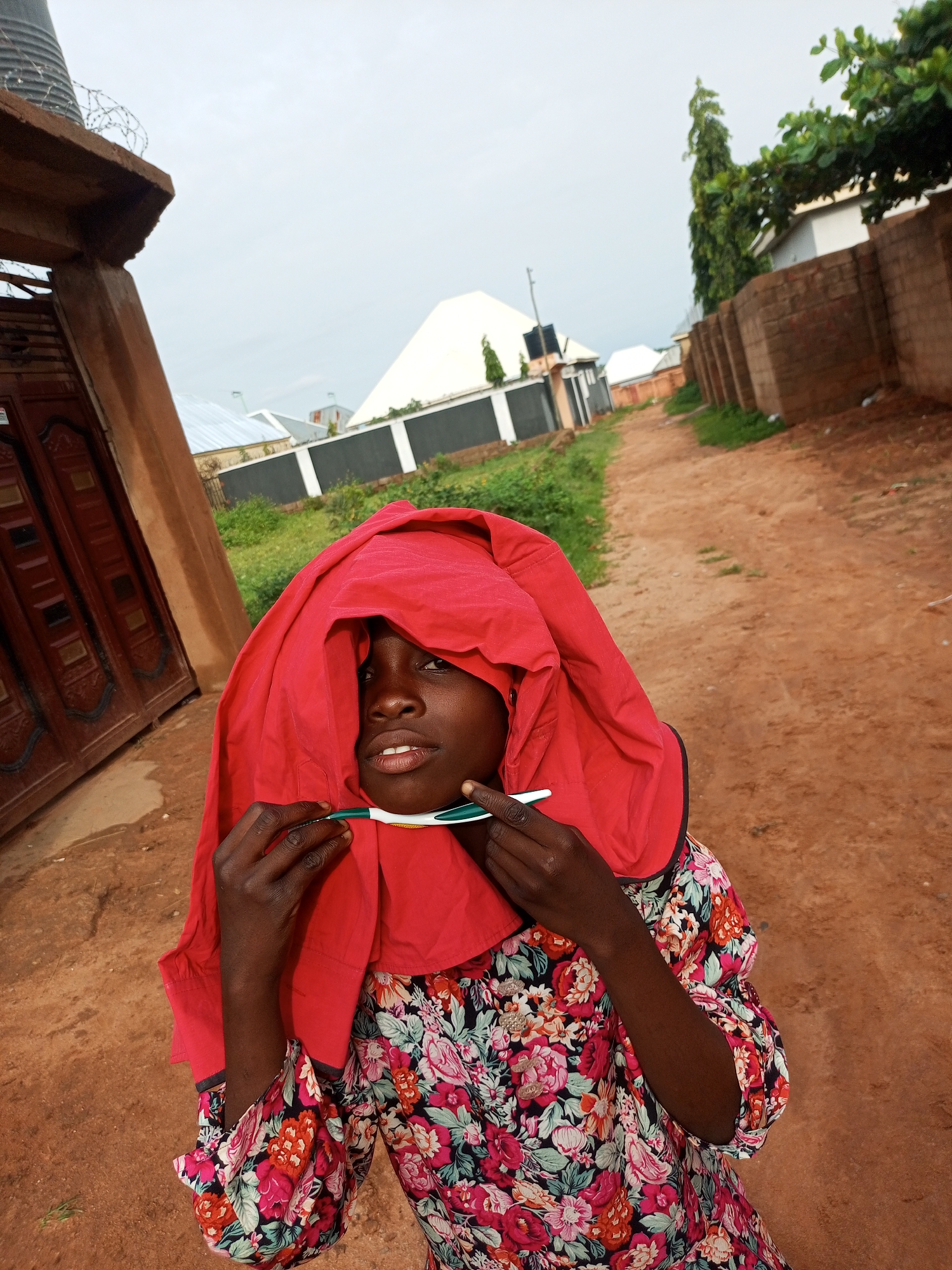 A young girl stands in a dirt road, holding a toothbrush up to her face, which is partially covered by a red cloth draped over her head. She wears a colorful floral dress. In the background, there is a brick wall and houses with white roofs, suggesting a residential area in the city of Tempure, Nigeria. The sky is overcast.