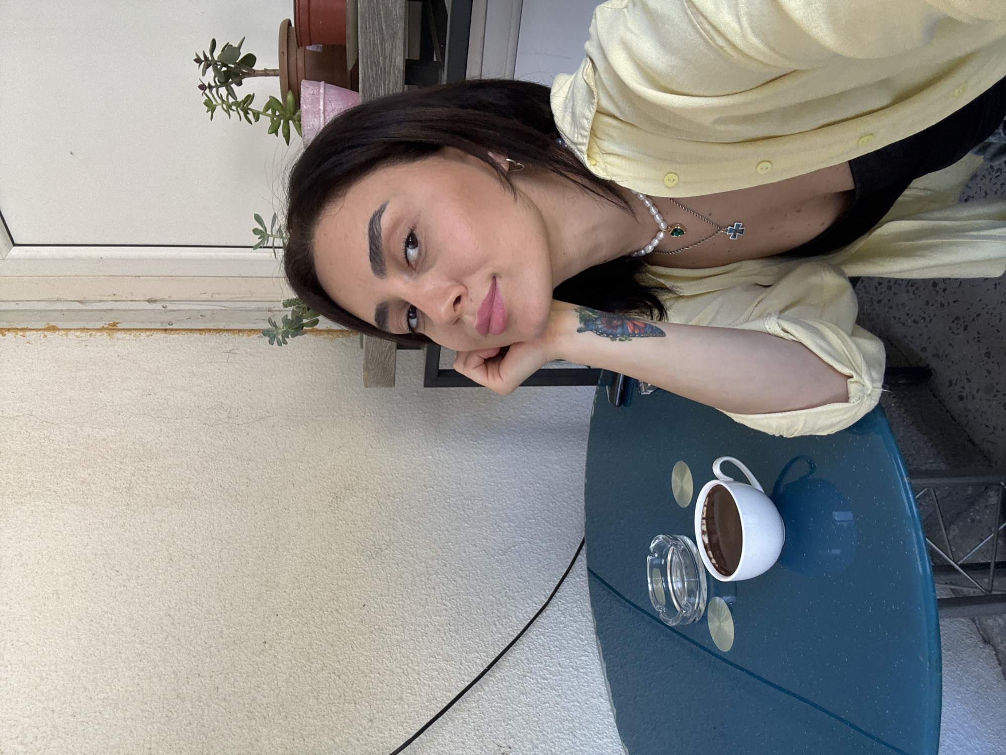 A medium shot captures a woman sitting at a teal-colored table outdoors, with a light-colored stucco wall in the background. She has dark hair, fair skin, and is wearing a light yellow button-up shirt over a black top. A pearl necklace with a green pendant and a silver chain with a cross pendant adorn her neck. She is resting her chin on her hand, and a butterfly tattoo is visible on her left forearm. On the table in front of her sits a white mug filled with a dark beverage, a glass ashtray, and two round, metallic coasters. The scene appears to be in Tbilisi, Georgia, suggested by the style of the stucco wall and the presence of potted plants on a shelf in the background, common in outdoor cafe settings in the city.
