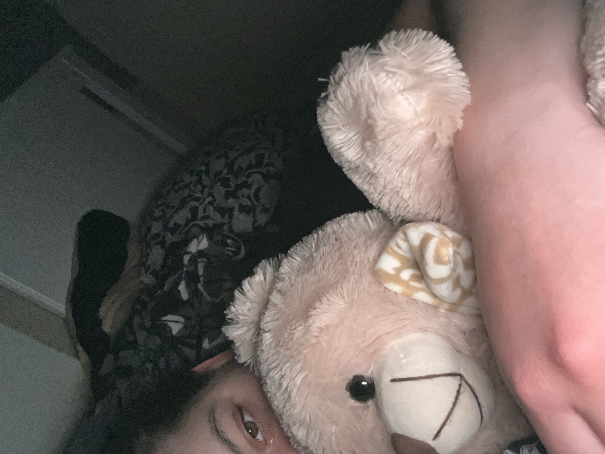 A person lies in bed, embracing a pink teddy bear with a patterned ear. The person's face, with one visible eye, is nestled near the bear. Behind them is a patterned blanket and a partial view of a doorframe in a dimly lit room. The location is unknown and cannot be determined to be Plovdiv, Bulgaria from the given image.