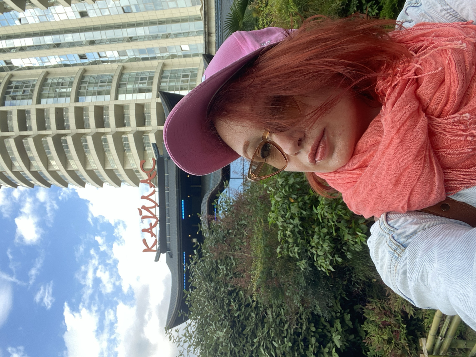 The photograph depicts a woman with red hair, wearing a pink baseball cap, brown-framed glasses, and a coral-colored scarf, likely in Sochi, Russia. The background includes a modern, high-rise building with a curved facade, as well as greenery and a structure with the name "KAMIKAZE" in red letters. The sky is visible with white clouds, and the overall setting suggests an outdoor urban environment. The woman appears to be posing for the camera. She is also wearing a light-blue jacket, the picture seems to be taken during daylight.