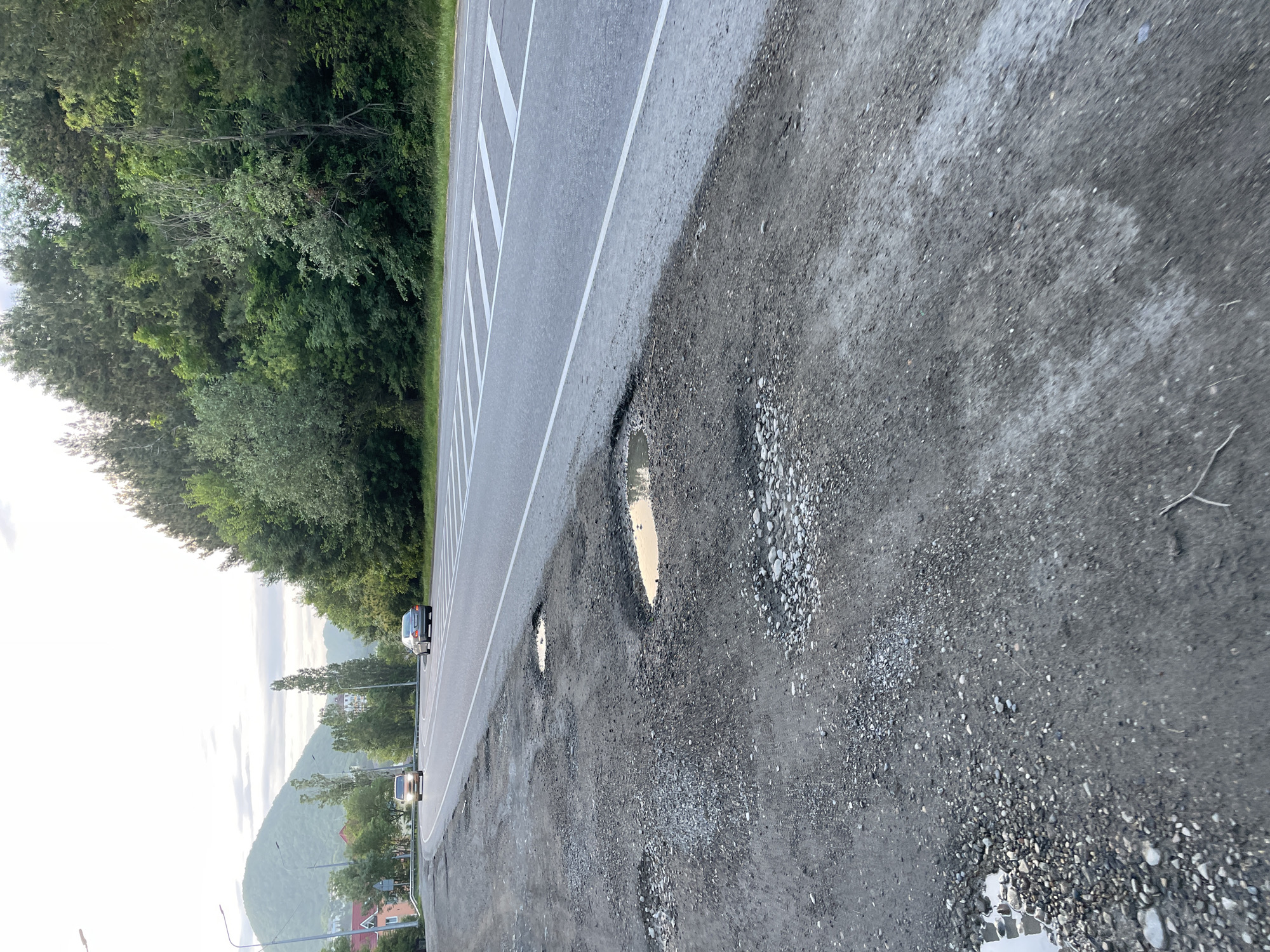 The image depicts a road in Olginka, Russia, that is in disrepair. The road surface is cracked and has several potholes, some of which are filled with water. The road has white lines indicating lane markings. A strip of trees and vegetation runs alongside the road. In the distance, a mountain and some buildings are visible. Two vehicles are seen on the road. The sky appears overcast. The general impression is of a road in poor condition in a semi-urban, hilly landscape.