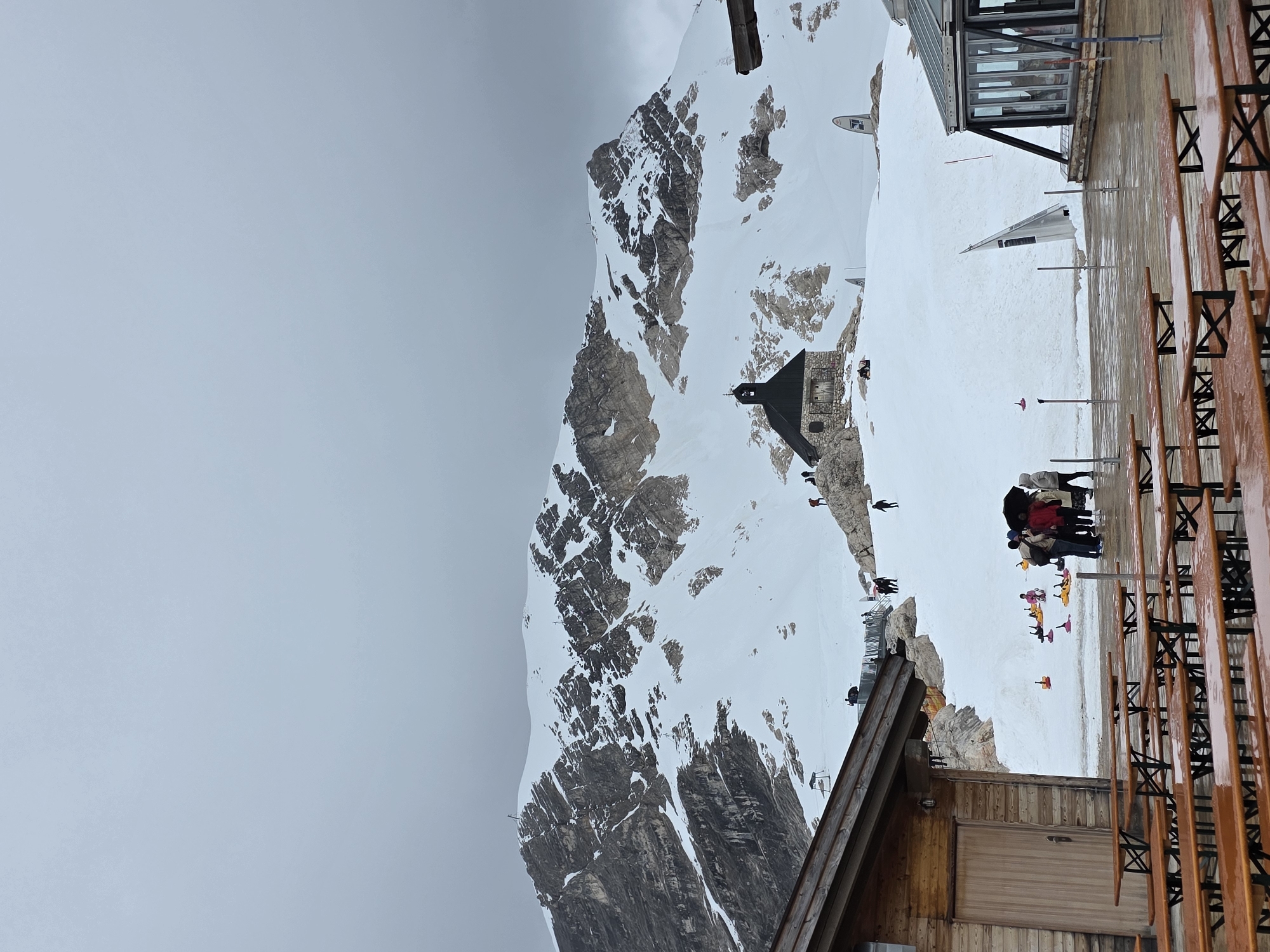 The image depicts an outdoor, high-altitude mountain scene characterized by substantial snow cover and visitor infrastructure, consistent with an Alpine resort plateau. In the immediate foreground, a wooden deck hosts multiple wet, empty wooden picnic tables and benches. To the left, a section of a rustic wooden building with a gabled roof and a closed wooden door is visible. To the right, a modern building with large glass windows and a dark roof, likely a restaurant or viewing area, is partially shown.
Beyond the deck, a broad expanse of snow-covered ground features scattered individuals and small groups of people, dressed in various cool-weather jackets; one person holds an open black umbrella. Several brightly colored plastic sleds, predominantly yellow, pink, and purple, are visible on the snow, indicating recreational activities. In the midground, a small stone chapel with a dark, steep gabled roof and a bell tower is centrally situated on a rocky outcrop amidst the snow. Various white and black markers or signs are intermittently spaced across the snow-covered area.
The background is dominated by a majestic, rugged, snow-capped mountain range, revealing distinct dark rock formations interspersed with snow. The sky above is uniformly overcast and grey. Small cross-like structures are discernible atop some of the distant peaks.