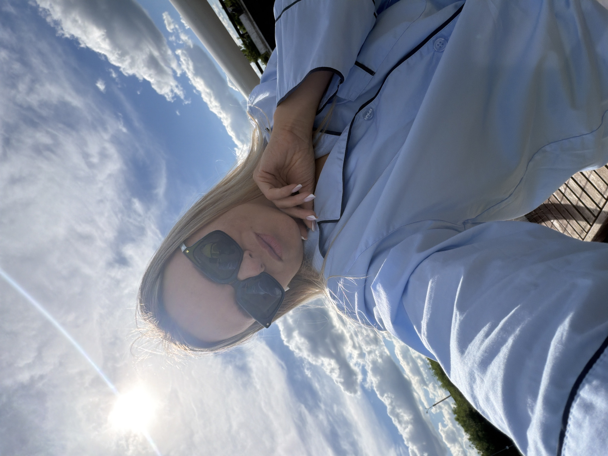 A blonde woman takes a selfie outdoors in Votkinsk, Russia, captured from a slightly low angle and tilted perspective. She is seen from the chest up, wearing a light blue, long-sleeved shirt with black piping along the placket, collar, and cuffs. She has long, straight blonde hair and is wearing stylish, square-framed dark sunglasses. Her left hand is raised towards her face, with fingers lightly touching her cheek, revealing manicured nails with white tips.
The setting is an bright outdoor environment under a vast, partly cloudy blue sky. Various types of white clouds, including cumulus and cirrus, are scattered across the frame, indicating pleasant weather. The sun is visible in the lower-left portion of the image, casting a strong lens flare that illuminates parts of the scene. In the background, to the left, a tall white pillar or structure is visible, and on the right, a wooden railing or deck can be seen, suggesting a balcony, terrace, or elevated outdoor area. Distant greenery, likely trees, is also visible under the horizon line. The time of day appears to be daytime, given the bright sunlight. No visible text is present in the image. The overall mood is casual and relaxed.