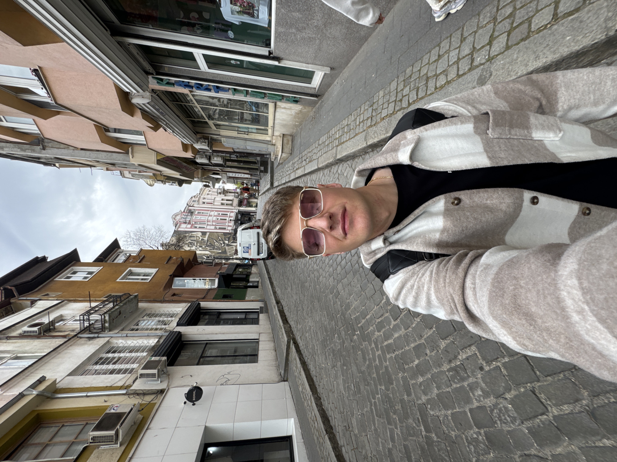 An outdoor selfie captures a young man on a cobblestone street in what appears to be an urban area, likely Plovdiv, Bulgaria, given the architectural style and hilly background. The image is rotated 90 degrees clockwise.
The main subject is a light-skinned man, seen from the chest up, with short, light brown hair styled to the side. He is wearing large sunglasses with pink-tinted lenses, a black t-shirt, and a loose-fitting jacket with a beige and white checkered or plaid pattern. A thin gold chain is visible around his neck, and a black bag strap crosses his chest. He has a neutral to slightly smiling expression, looking directly towards the camera.
The background reveals a narrow street flanked by multi-story, older buildings with diverse facades, some painted in earthy tones like ochre and brown. Many windows, air conditioning units, and a small metal balcony are visible on the buildings to the left. To the right, there are shopfronts with display windows, one of which showcases a white t-shirt with a floral graphic. The street itself is paved with dark, irregularly shaped cobblestones. In the distance, a white van is parked, and more buildings climb up a hillside, suggesting a historic or hilly district. The sky is overcast, indicating a cloudy daytime.
Visible text includes "BORBAL" on a white sign below one building. On the white van's license plate, a European format is visible, possibly "BG" for Bulgaria, along with characters such as "F 2769 CT." Other shop signs have indistinct text. Another person's feet in sneakers can be seen in the upper right corner of the original image, implying they are walking alongside the subject.