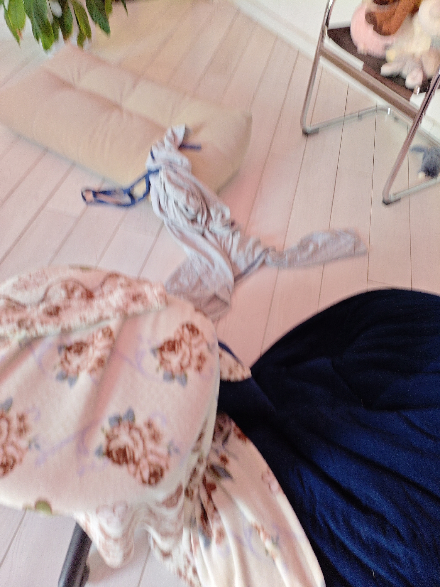 The image depicts an indoor scene with a light-colored wooden floor. In the foreground, a large, dark blue fabric is spread out. To the left of this, a plush blanket with a floral pattern of roses and blue ribbons is draped over a dark object, possibly a chair or stool. A light-colored mattress or futon is visible further back. Near the futon, a light gray garment, possibly a t-shirt or sweatshirt, is draped, with blue straps or ties attached. In the background, a metal chair with a dark seat and a light-colored object on it, possibly stuffed animals, is visible. The overall impression is of a domestic setting that appears somewhat untidy or in the process of being organized. The location is identified as Трудовское сельское поселение, Ukraine.
