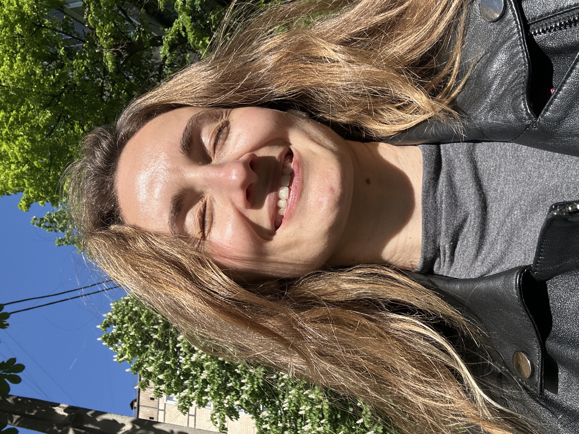 A woman with long, blonde hair is smiling broadly with her eyes closed. She is wearing a gray t-shirt and a black leather jacket. The background features green trees and a blue sky. A building is partially visible at the bottom left of the frame. The location is outdoors, likely in a park or residential area. I have no information about whether the image was taken in Kryvyi Rih, Ukraine.
