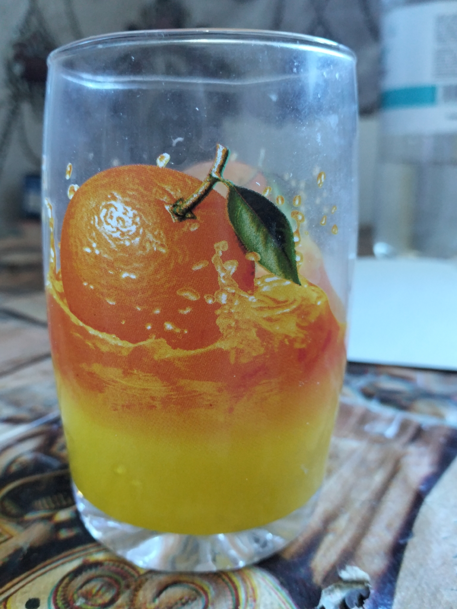 A close-up, eye-level shot shows a clear glass filled with a yellow and orange liquid, likely juice, with a printed image of an orange slice and leaf wrapped around the outside of the glass. The orange is depicted in a splash of liquid, creating a dynamic visual. The glass is positioned on a surface with an abstract, colorful pattern, possibly a tablecloth or placemat. In the background, a blurred white object with a teal band is visible, suggesting it might be a bottle or container. The lighting is soft, with some highlights and shadows, and the overall scene is indoors. The location of Kogon City, Uzbekistan, is not discernible from the visual elements present in the image.