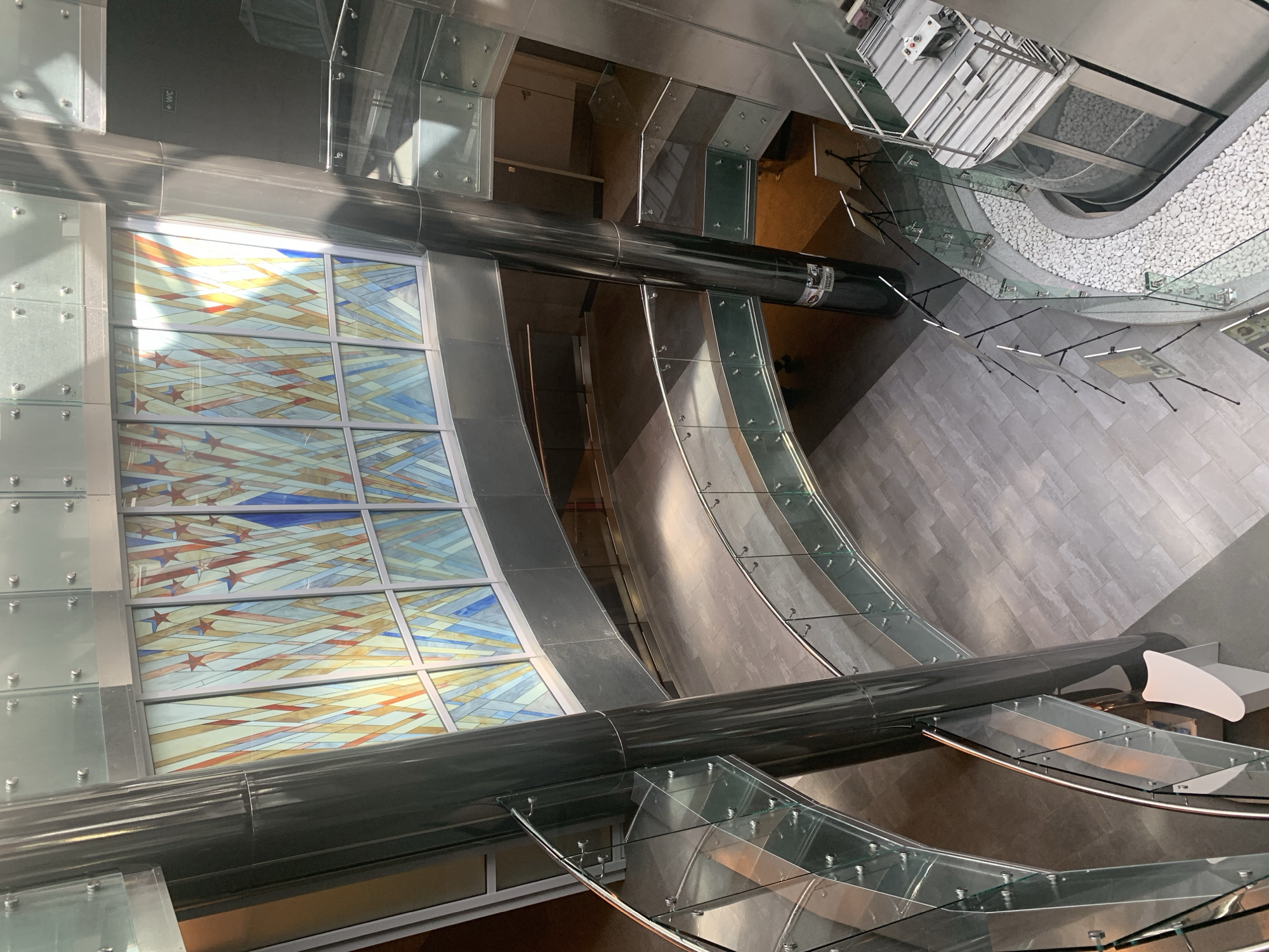 Here's a detailed description of the image:
**Location:** The image appears to be taken inside a modern architectural structure in Minsk, Belarus. This is based on the design elements and materials visible.
**Scene Description:**
The image presents an unusual perspective, likely from an elevated position looking down into the building's interior. The main features include:
* **Stained Glass:** A large, multi-paneled stained glass window dominates one side of the image. It features geometric patterns with a combination of colors, including blue, orange, and red. Some panels include a pattern of stars.
* **Architectural Structure:** The building's interior features curving walls, with elements that appear to be glass railings or partitions.
* **Metallic Components:** A prominent, large-diameter metallic pipe or structural element runs horizontally across the image. It appears to be attached to the building's framework.
* **Stairs/Escalators:** There are structures resembling stairs or escalators partially visible in the background of the image. Some are enclosed with glass.
* **Flooring:** The floor is tiled with light grey rectangular tiles.
* **Pebble area:** At the top of the image there's a section filled with many white pebbles.
* **Lighting:** The lighting is generally bright. It appears to be primarily from natural light sources coming through the glass elements of the building.
* **Other Details:** Small objects appear to be placed on the floor.
**Overall Impression:**
The image captures a modern and aesthetically driven interior space with a blend of natural light, architectural details, and artistic elements such as the stained glass.