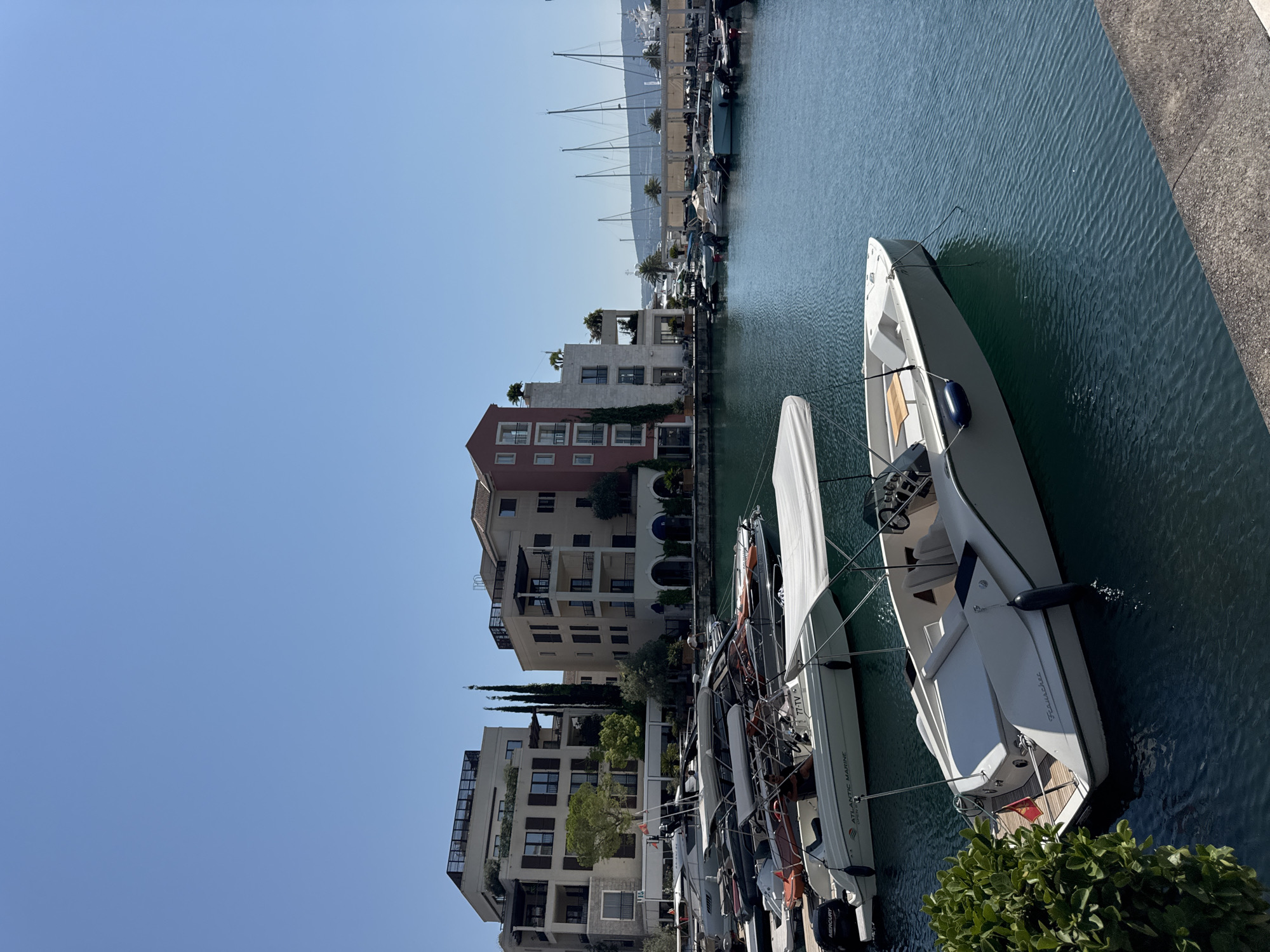 This is a daytime, eye-level, outdoor, wide-angle shot taken in Tivat, Montenegro. The image depicts a marina scene with several boats docked in the foreground and midground. The water is a vibrant turquoise color and appears calm with small ripples. In the background, there are modern multi-story buildings with balconies and windows, characteristic of waterfront architecture in Porto Montenegro. There are also several sailboats with tall masts visible further out in the harbor, suggesting a larger marina. The sky is clear and blue, indicating fair weather. The scene is serene and suggests a leisurely, upscale environment typical of a yachting hub.
