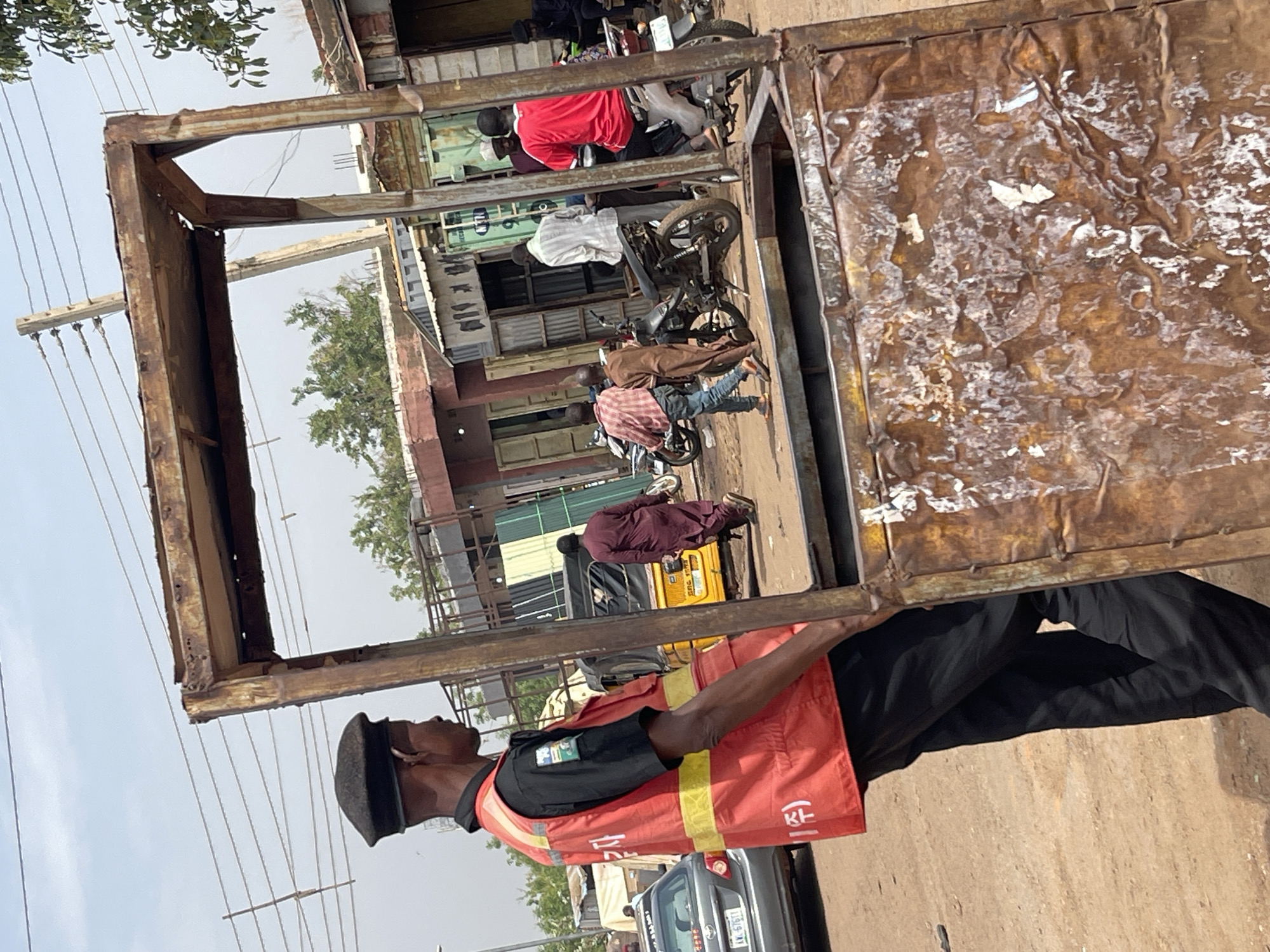 A person in a black cap, black shirt, black pants, and a bright red safety vest with vertical yellow stripes and white lettering is carrying a rusty metal frame. The frame is box-shaped, open on the viewer's side, with a sheet of heavily distressed metal forming the far side. The person appears to be walking along a street in Zaria, Nigeria. Behind them, motorcycles are parked along the sidewalk. People are visible, some on the motorcycles. A yellow tricycle vehicle is also visible. Cars and street lights line the street. Buildings form the backdrop. The sky is overcast.