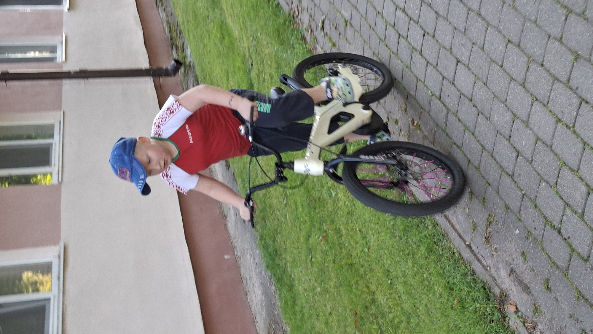 A young boy wearing a blue baseball cap and a red and white t-shirt is riding a cream-colored bicycle on a paved walkway. The boy is facing right, with his hands on the handlebars. The bicycle has black tires with pink spokes on the front wheel. The walkway is made of gray cobblestones and is bordered by a grassy area. In the background, there is a building with windows and a downspout. The scene appears to be in a residential area, possibly in Myadzyel, Belarus, given the context of Myadzyel, Belarus being a city.