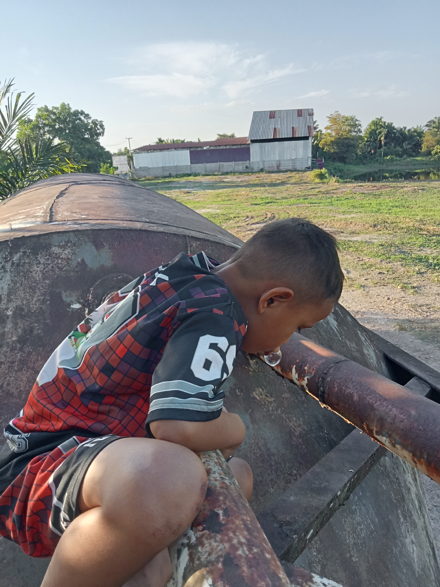 A young boy in a black and red patterned shirt with the number "6" visible on it is sitting on a rusty, curved metal object, possibly part of a derelict vehicle or structure. He is leaning forward, with his head down, and appears to be blowing a bubble, which is visible near his mouth. His shorts are also red and black with a similar pattern. The background shows a grassy field with some dirt tracks, and in the distance, there are buildings with metal roofs, trees, and a body of water. The sky is a clear blue with some faint white clouds. The lighting suggests it is late afternoon, with the sun casting long shadows. The overall impression is one of casual play in an outdoor, somewhat industrial or rural setting. The location is Kualu, Indonesia.