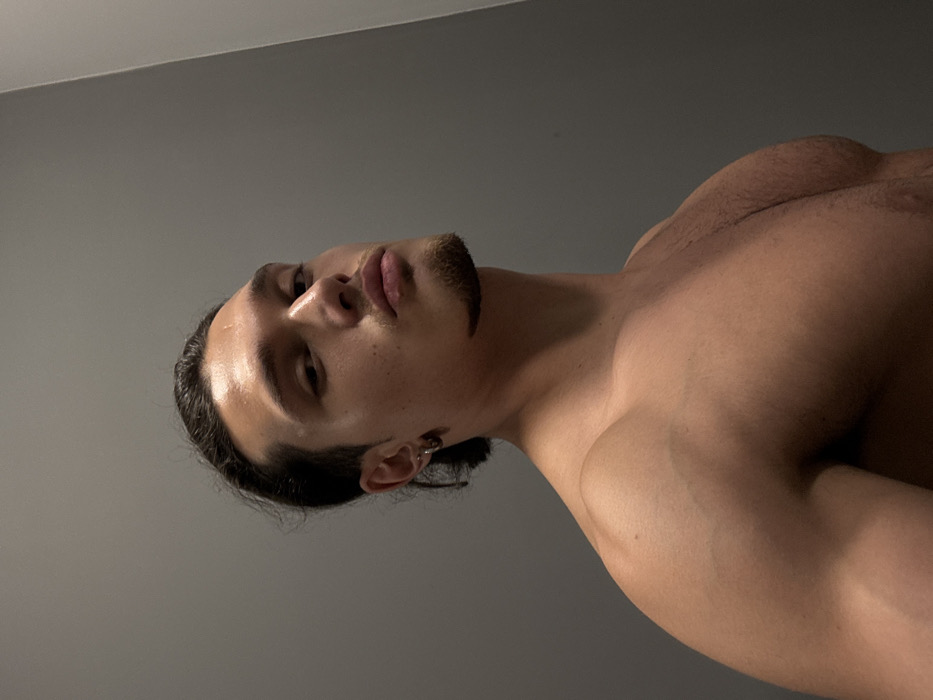 A young adult male, shirtless, is depicted in an indoor setting within Sofia, Bulgaria. He has a muscular physique, dark hair pulled back, a goatee and mustache, and multiple silver-toned hoop earrings in his left earlobe. He gazes directly towards the camera with a serious expression. The background consists of a plain, uniform gray wall, providing no additional environmental details or objects.