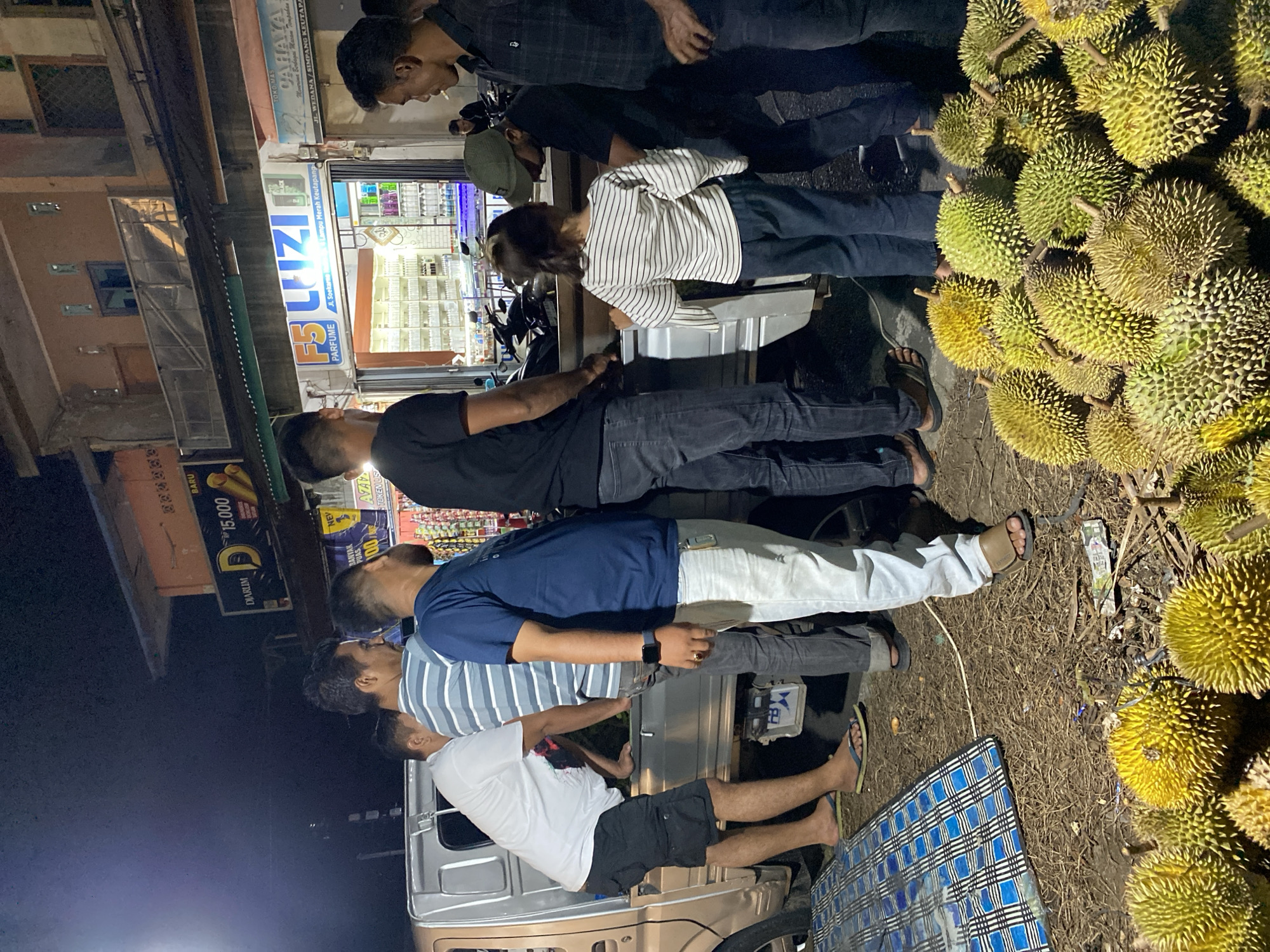 This media file captures a nocturnal scene in Lambheu, Indonesia, where a group of people are gathered around a stall or vehicle laden with durians. The primary subjects are the individuals, whose faces are mostly obscured or turned away, engaged in apparent interaction with the durian seller or examining the fruit. The setting appears to be a roadside stall or a temporary setup, indicated by the dimly lit background featuring storefronts with signage and displays of goods.
The prominent objects in the scene are the numerous durians, piled high and clearly visible due to their distinctive spiky exteriors. A vehicle, likely a truck or van, is partially visible, suggesting a mobile stall or a mode of transport for the fruit. Several people are standing and observing the durians. One person in the foreground is wearing a blue t-shirt and jeans, while another in a white t-shirt and black shorts is also visible.
The time of day is clearly night, given the darkness of the surroundings and the artificial lighting. The weather is not discernible from the image. Among the visible text, "DIARIM", "15.000", "FS LUZIA", "PARFUME", "Lampu Merah Keutapang", "TOKO MAS CAHAYA", and "JL. WEDANA/SIMPANG KEUTAPA" are present on various signs, suggesting the commercial nature of the location. The overall mood appears casual and transactional, typical of a nighttime fruit market.