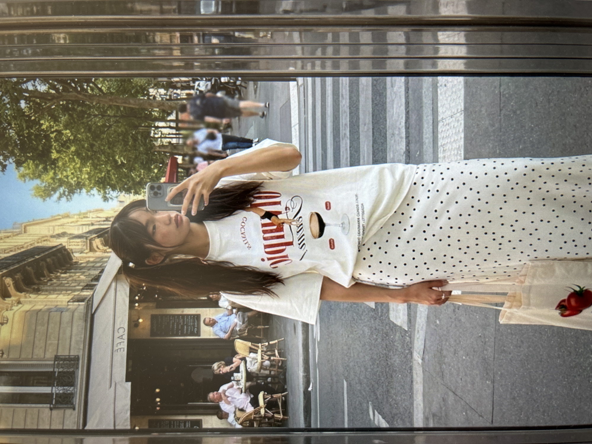 An Asian woman with dark hair tied in pigtails stands in front of a reflective surface, taking a selfie with her phone. She is wearing a white t-shirt with a graphic print and a long, white skirt with black polka dots. She is holding a canvas tote bag with a strawberry graphic. In the background, several people are seated at tables outside a cafe, and others are walking on a street with a crosswalk. The architecture in the background suggests a European city.