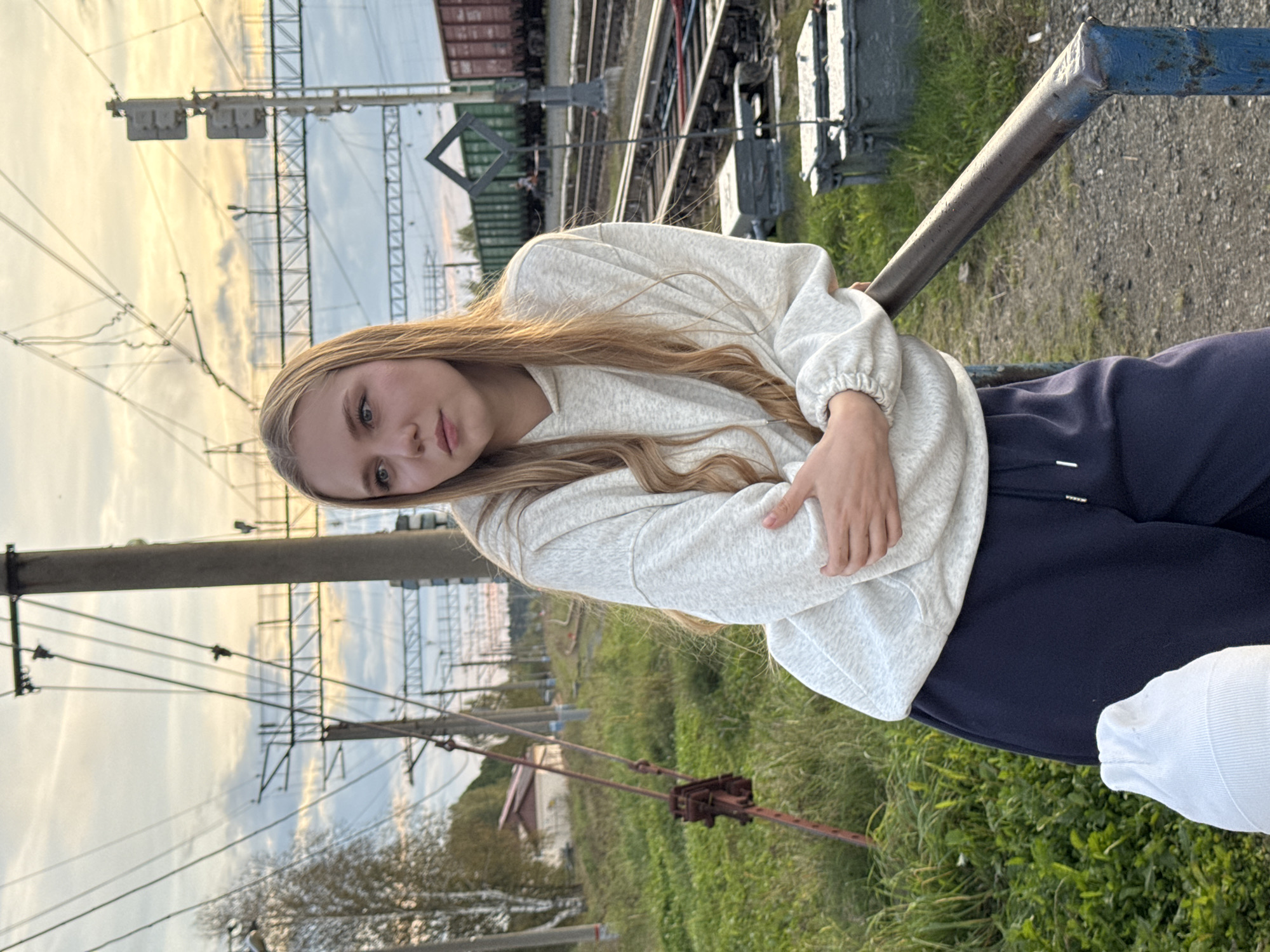 A young woman with blonde hair is standing by a metal railing next to a railway track in Комарихинский, Russia. She is wearing a light gray hooded sweatshirt and dark blue sweatpants. Her arms are crossed, and she is looking off to the side. In the background, there are train cars, power lines, and vegetation. The sky is cloudy with the sun setting, casting a warm glow.