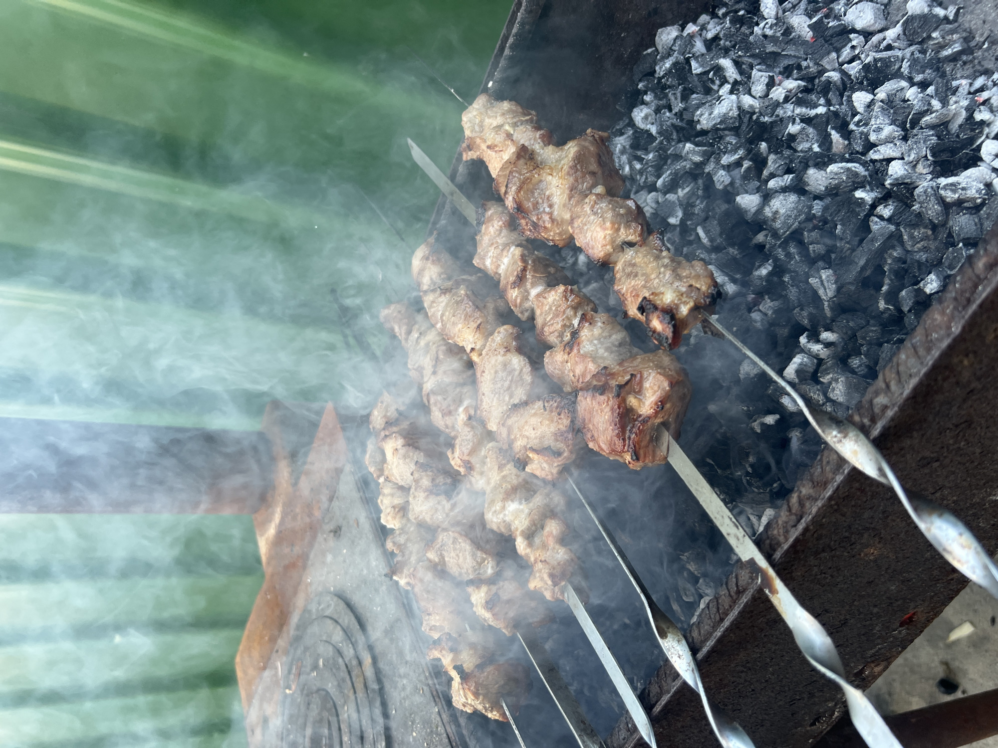 The image shows meat skewers being grilled over hot coals in a metal brazier. The skewers are positioned horizontally, with several pieces of meat threaded onto each one. Smoke billows upwards from the coals and the cooking meat, creating a hazy atmosphere. In the background, a green, striped surface is visible, possibly a fence or a curtain. The scene appears to be set outdoors, with a rustic stove or cooking apparatus to the left of the brazier. The overall impression is one of an outdoor barbecue or cooking event.