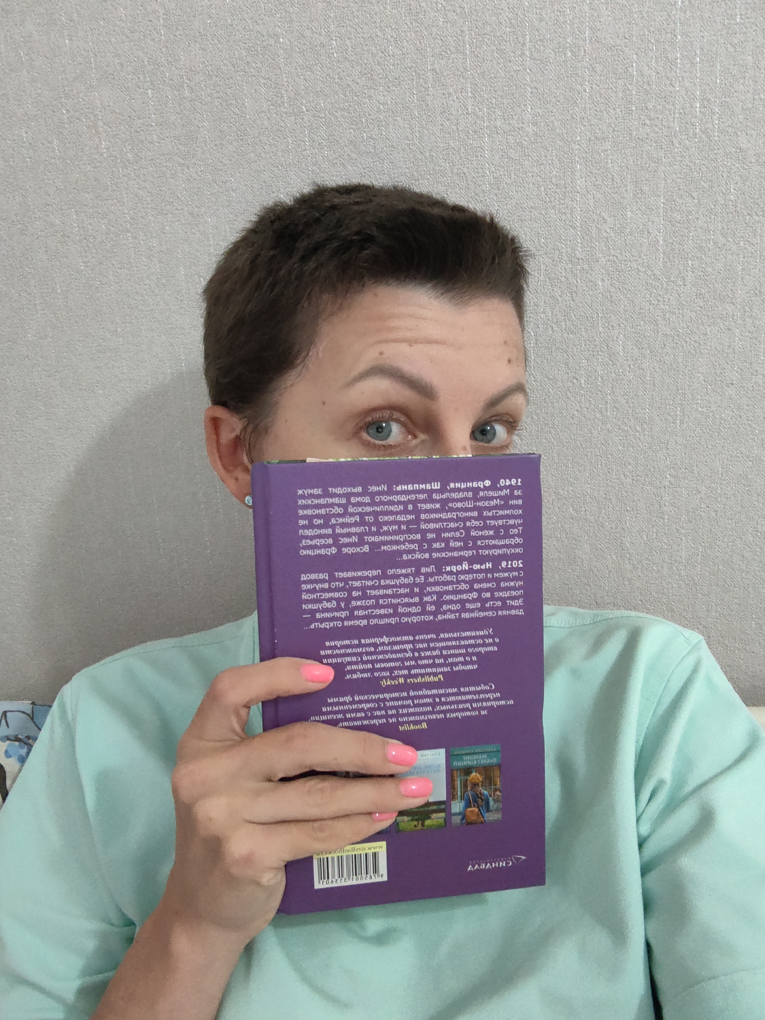 A young woman with short brown hair, wearing a teal t-shirt, is holding a purple book in front of her face, obscuring most of it. Her blue eyes are visible above the book. The book has text and images on it, including a barcode and a logo. The background is a textured gray wall. The image was taken indoors. There is no information available to determine the location in the city of Myadzyel, Belarus.