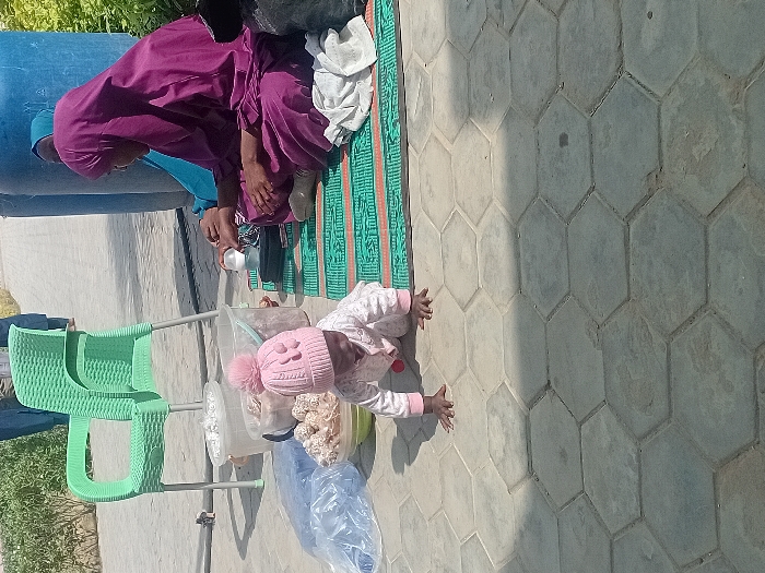 The scene is an outdoor setting in the city of Kwazailewa, Nigeria, under direct sunlight, indicated by strong shadows on the hexagonal grey concrete pavers. In the foreground, an infant is crawling on the pavement, dressed in a light-colored long-sleeved outfit and a pink knitted hat with a pom-pom. To the right, a woman in a dark purple hijab and matching long dress sits cross-legged on a green and orange patterned mat, holding a clear baby bottle with a white cap. A second woman, wearing a light blue hijab, is partially visible behind the first, also seated on the mat. Adjacent to the mat and the infant, an empty light green plastic chair with a woven pattern stands on the pavement. Near the chair are two translucent white plastic buckets, one containing unidentified light-colored contents, and a light-colored bowl filled with numerous small, spherical, light-colored items, possibly food products. A transparent blue plastic bag rests on the pavement near the bowl. Behind the seated women, two large blue plastic barrels are visible, partially obscured. In the distant background, two indistinct figures walk away from the viewer on what appears to be an unpaved area with some sparse green vegetation.